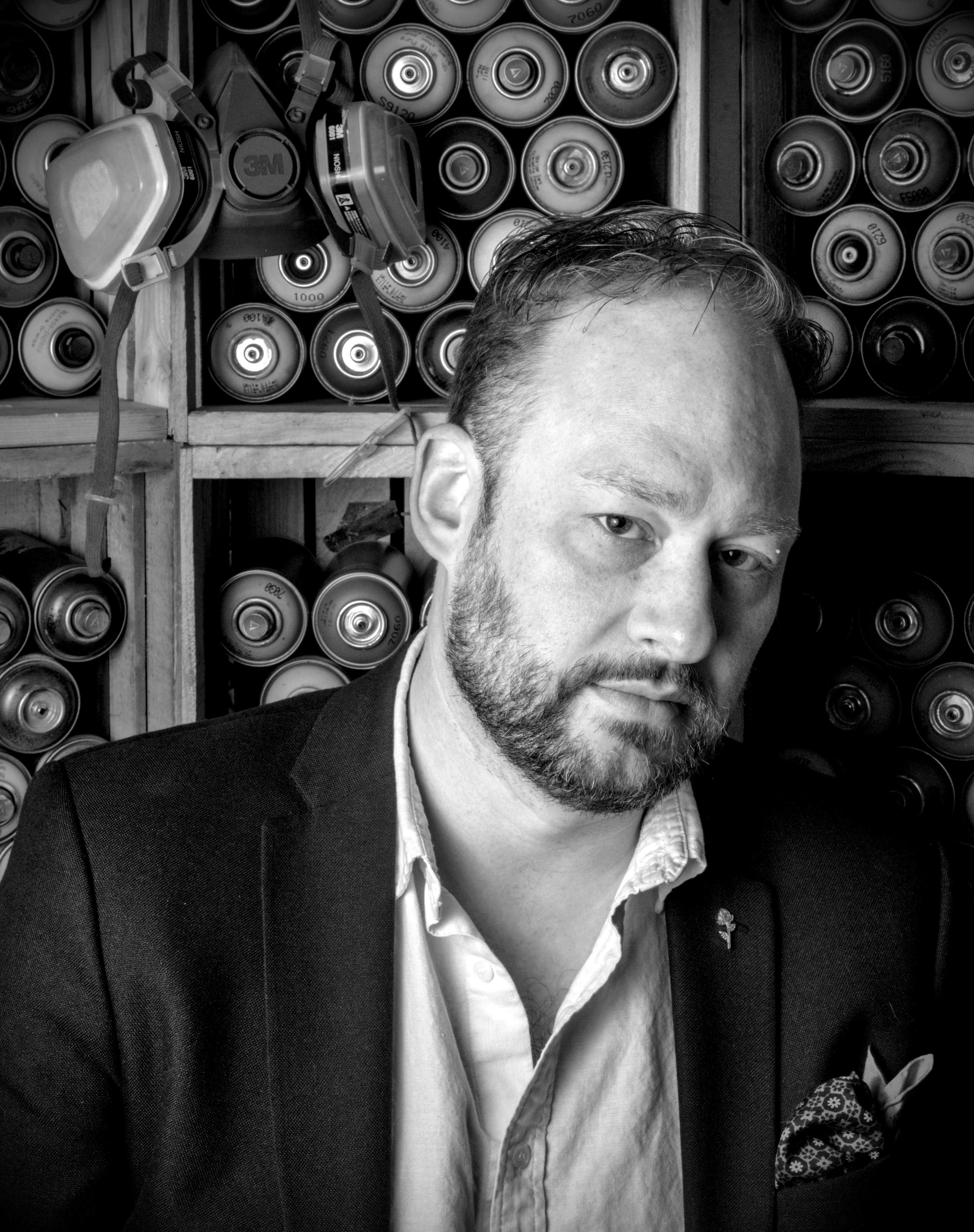 Bearded man in a dark blazer and open-collared shirt in front of shelves stacked with spray paint cans and respirator masks.