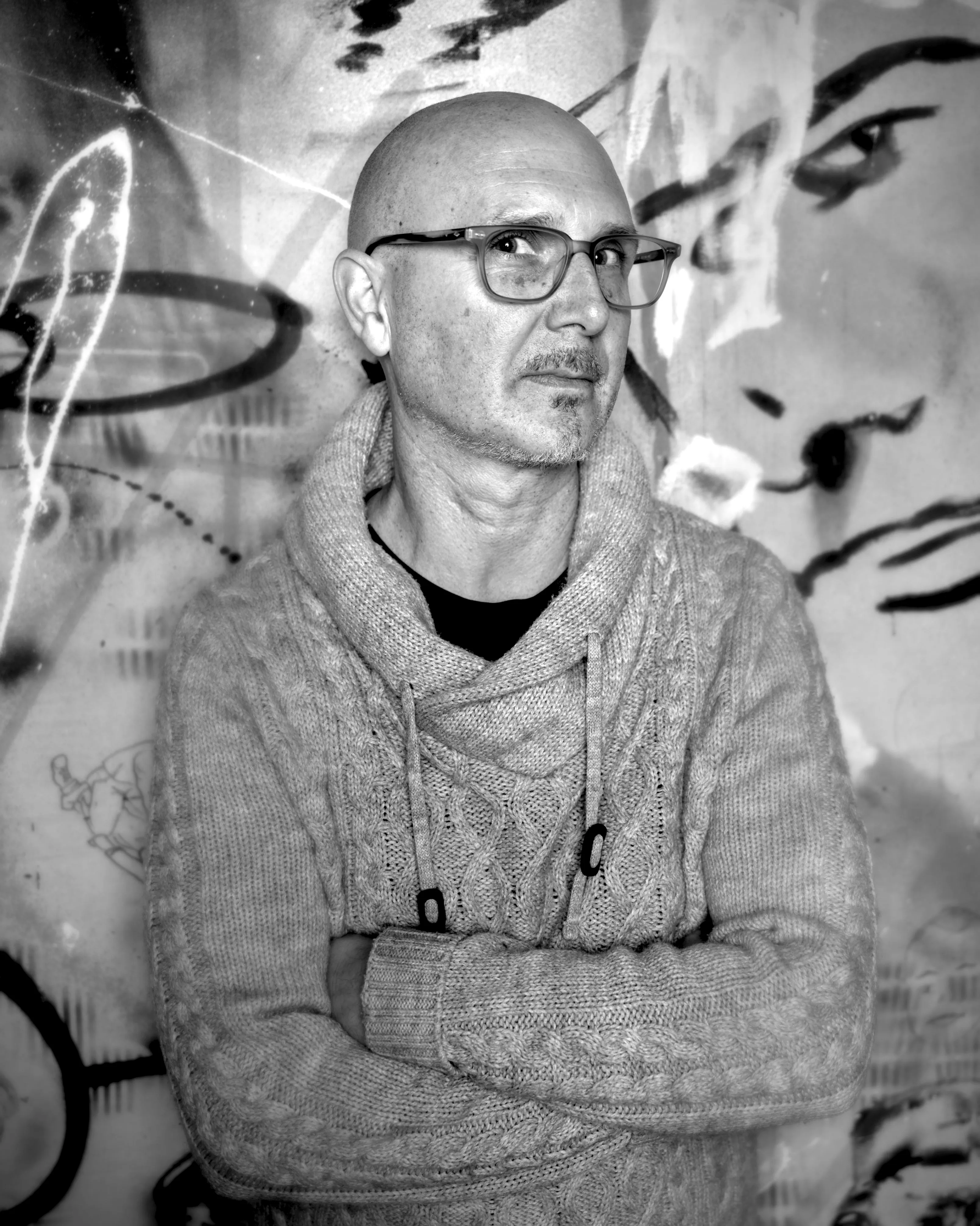 Bald man with glasses and a mustache, arms crossed, in a chunky knit sweater in front of a graffiti-covered wall.