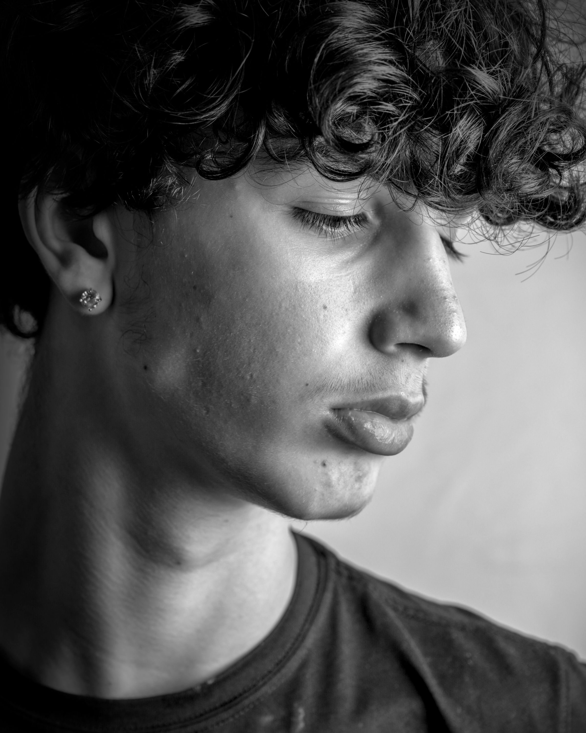 Young person with curly hair and a stud earring, shown in profile with eyes lowered, wearing a dark t-shirt.