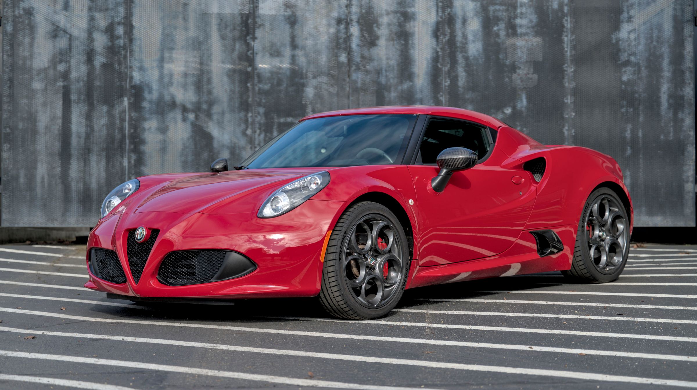 Red sports coupe with dark alloy wheels parked on a striped pavement in front of a weathered metal wall.