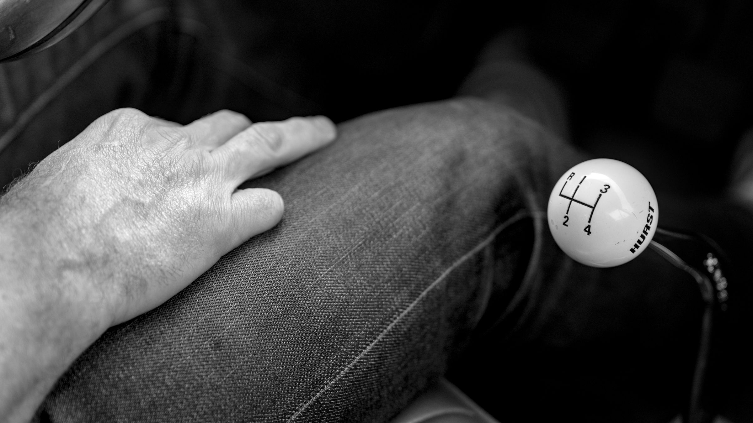Hand resting on a denim-clad knee beside a white Hurst gear shift knob with a four-speed pattern.