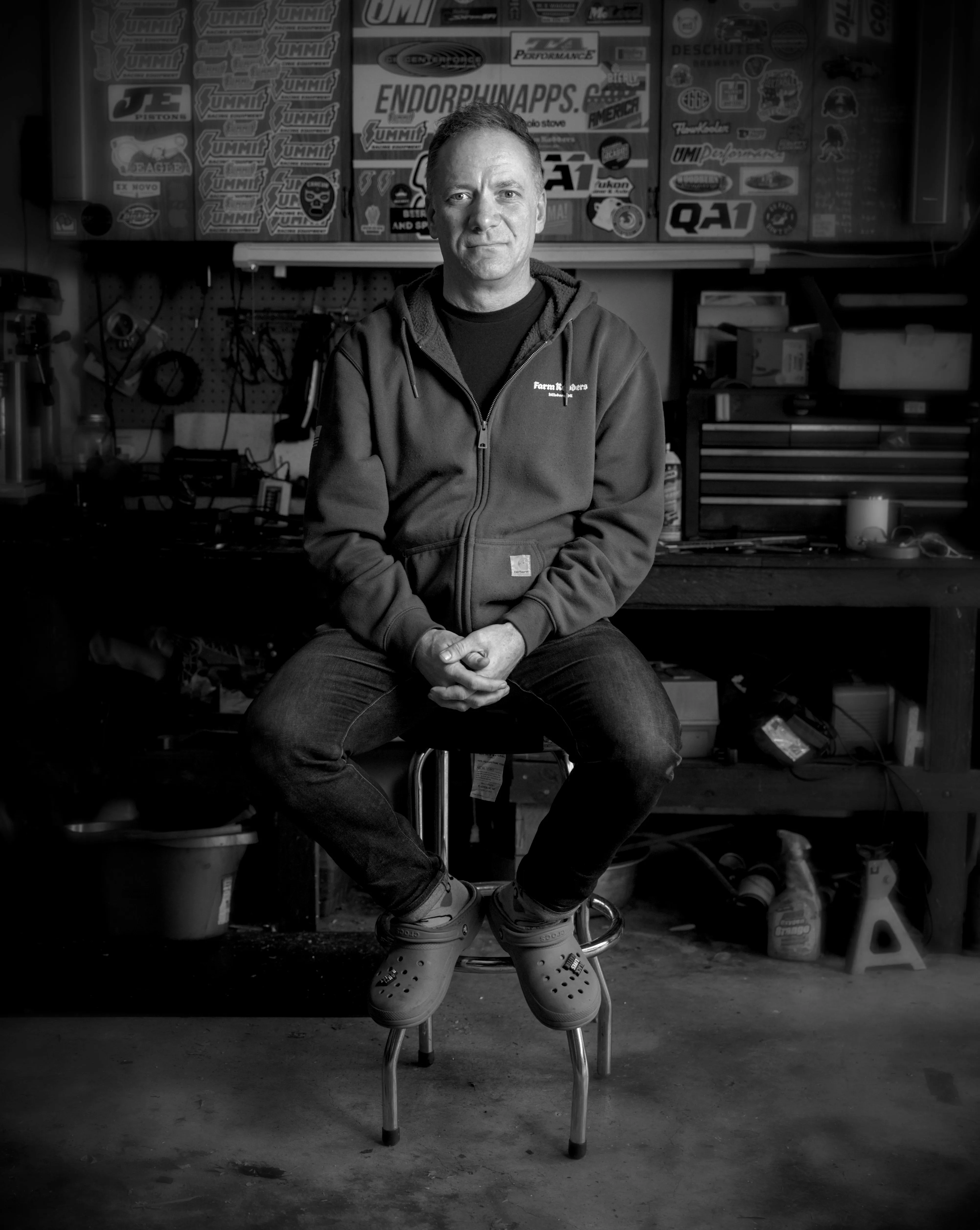 Man in a zip hoodie and clogs sits on a stool in a garage workshop with a sticker-covered panel behind him.