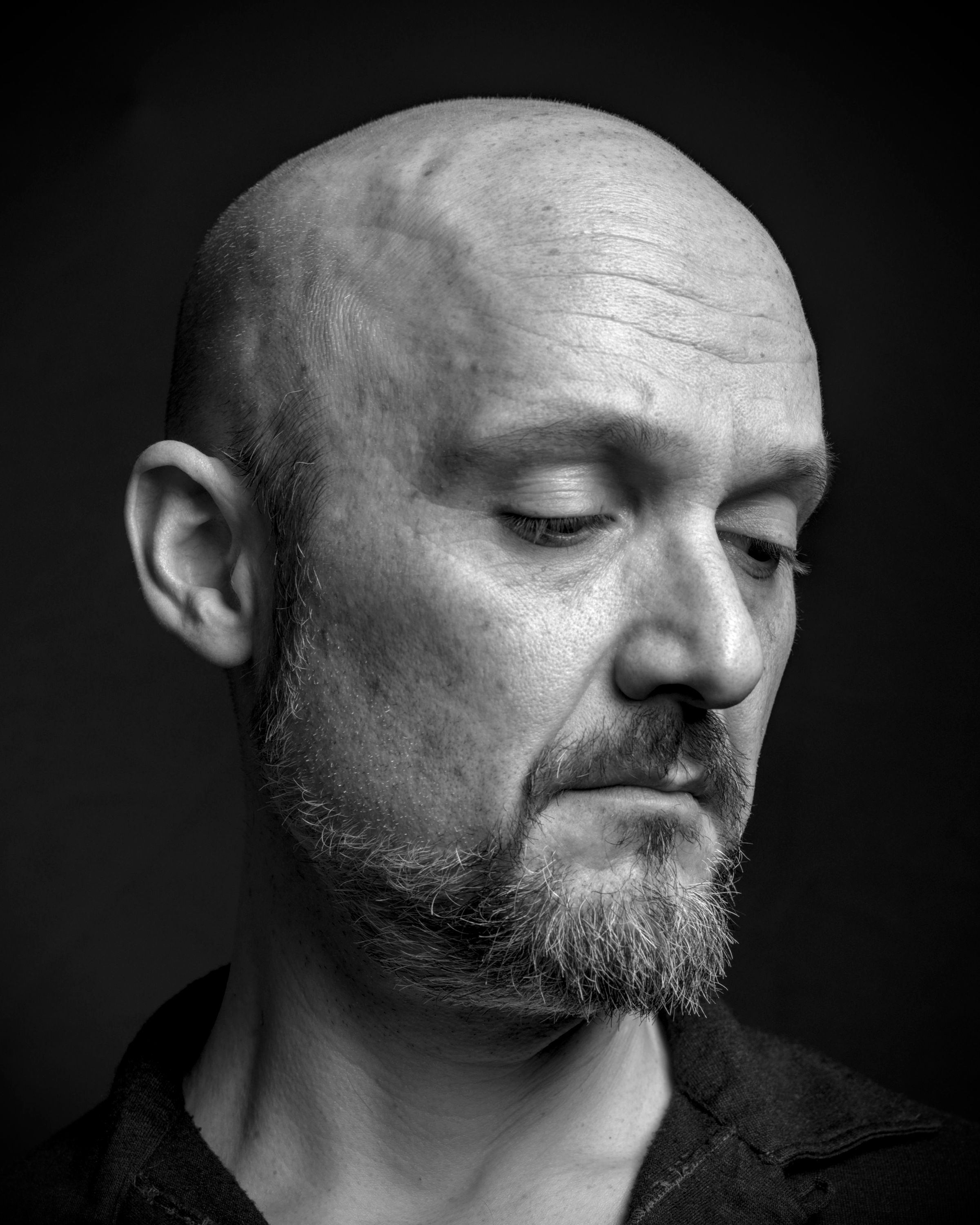 Close portrait of a bald, bearded man looking down against a dark background.