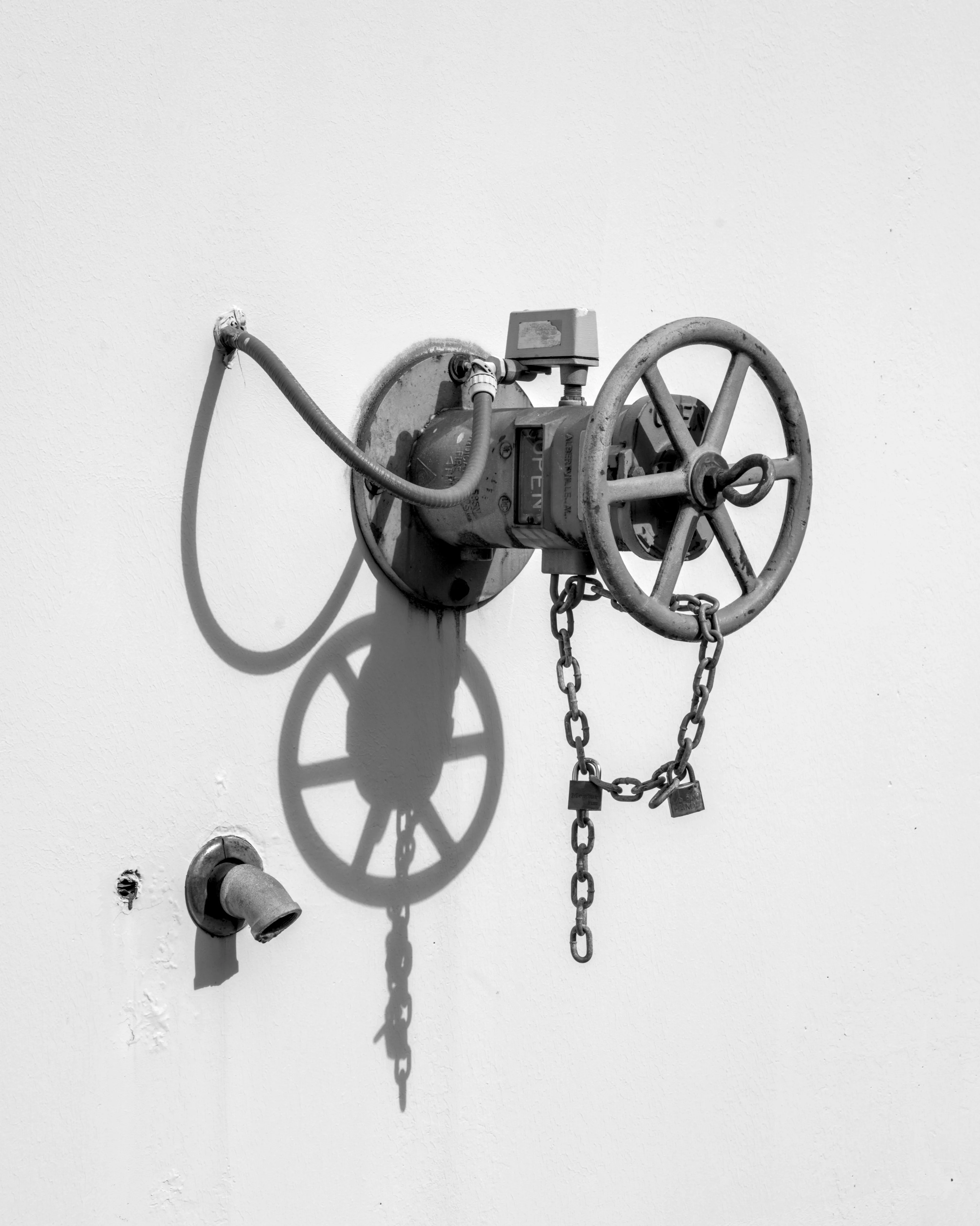 Wall-mounted valve with a spoked handwheel and padlocked chain casting shadows on a white wall.
