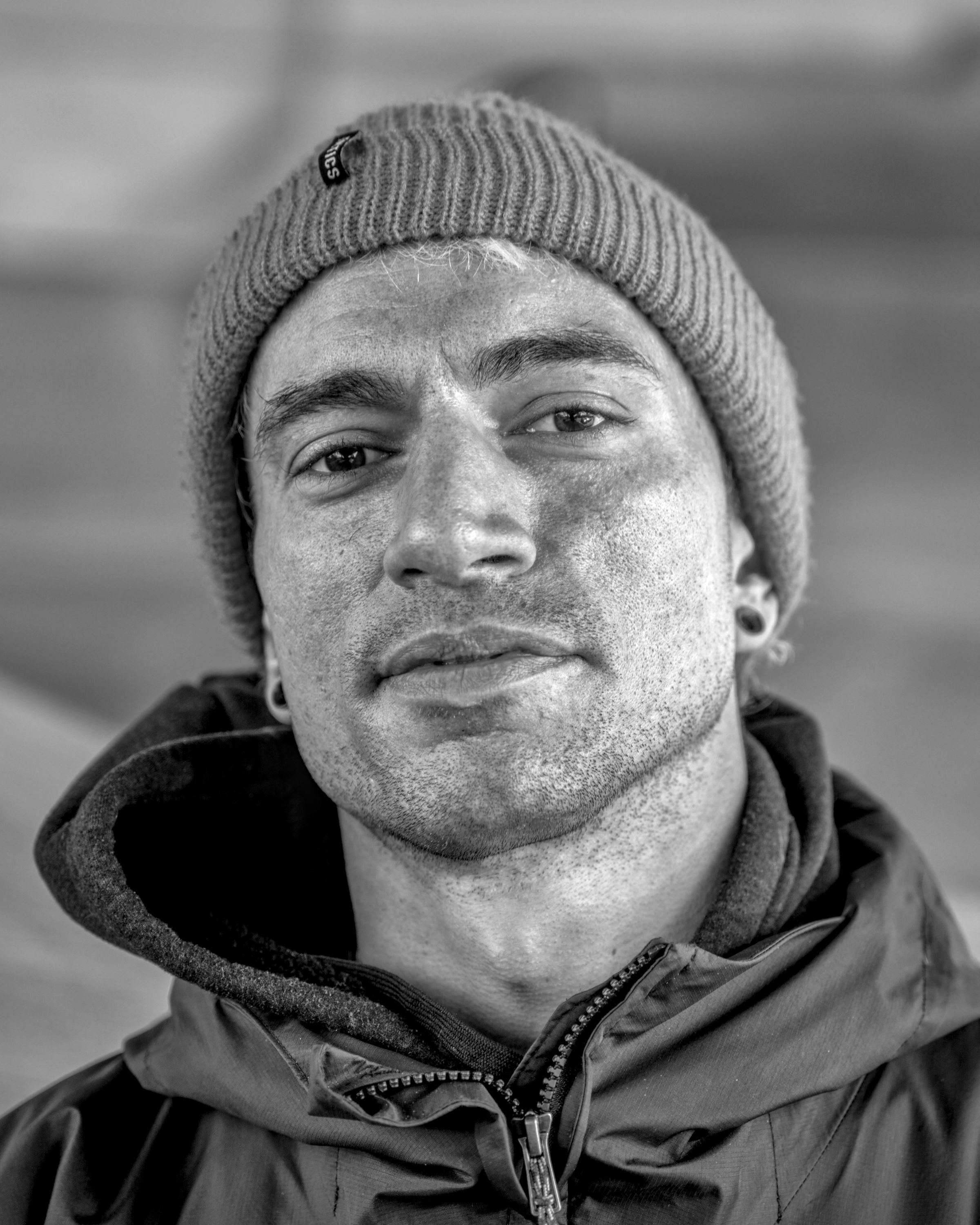 Close-up of a man with stubble in a knit beanie and a zipped hooded jacket, looking at the camera with a slight smile.