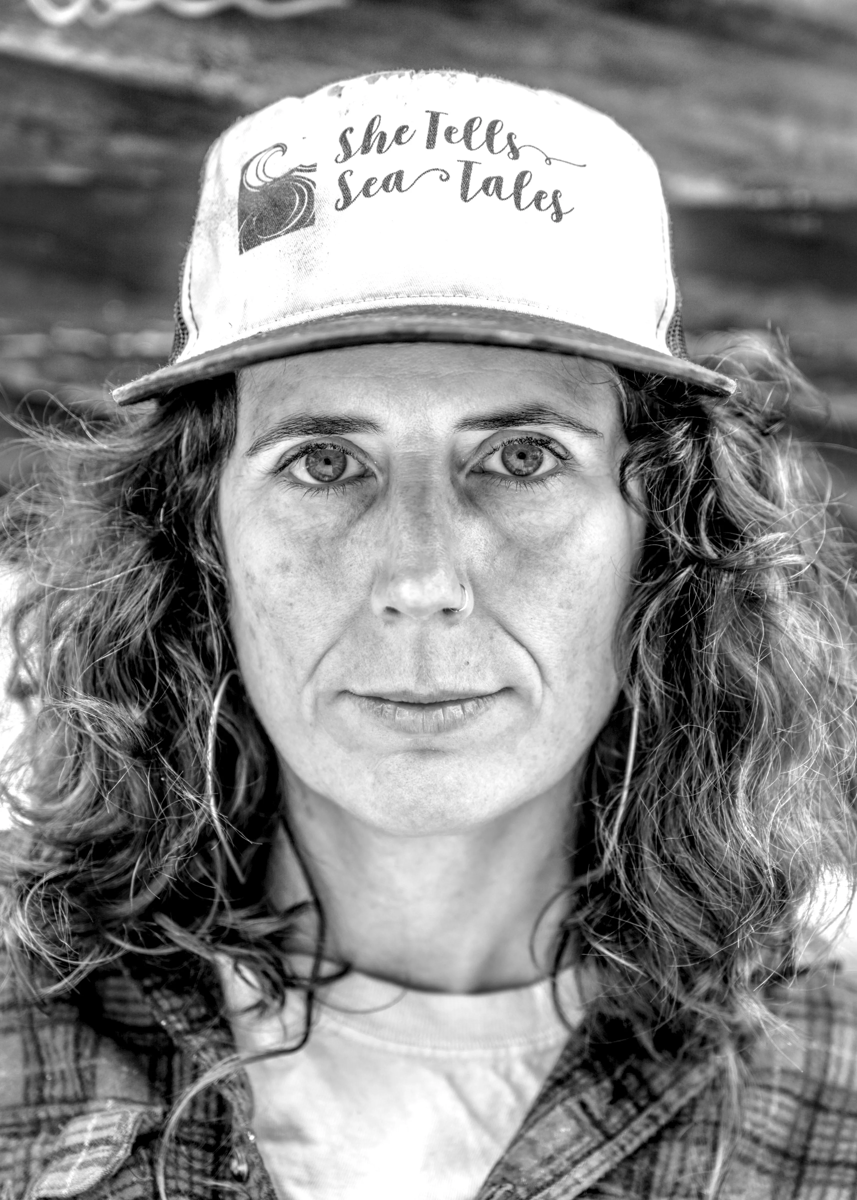 Close portrait of a person with wavy shoulder-length hair wearing a light trucker cap reading 'She Tells Sea Tales' and a plaid shirt.