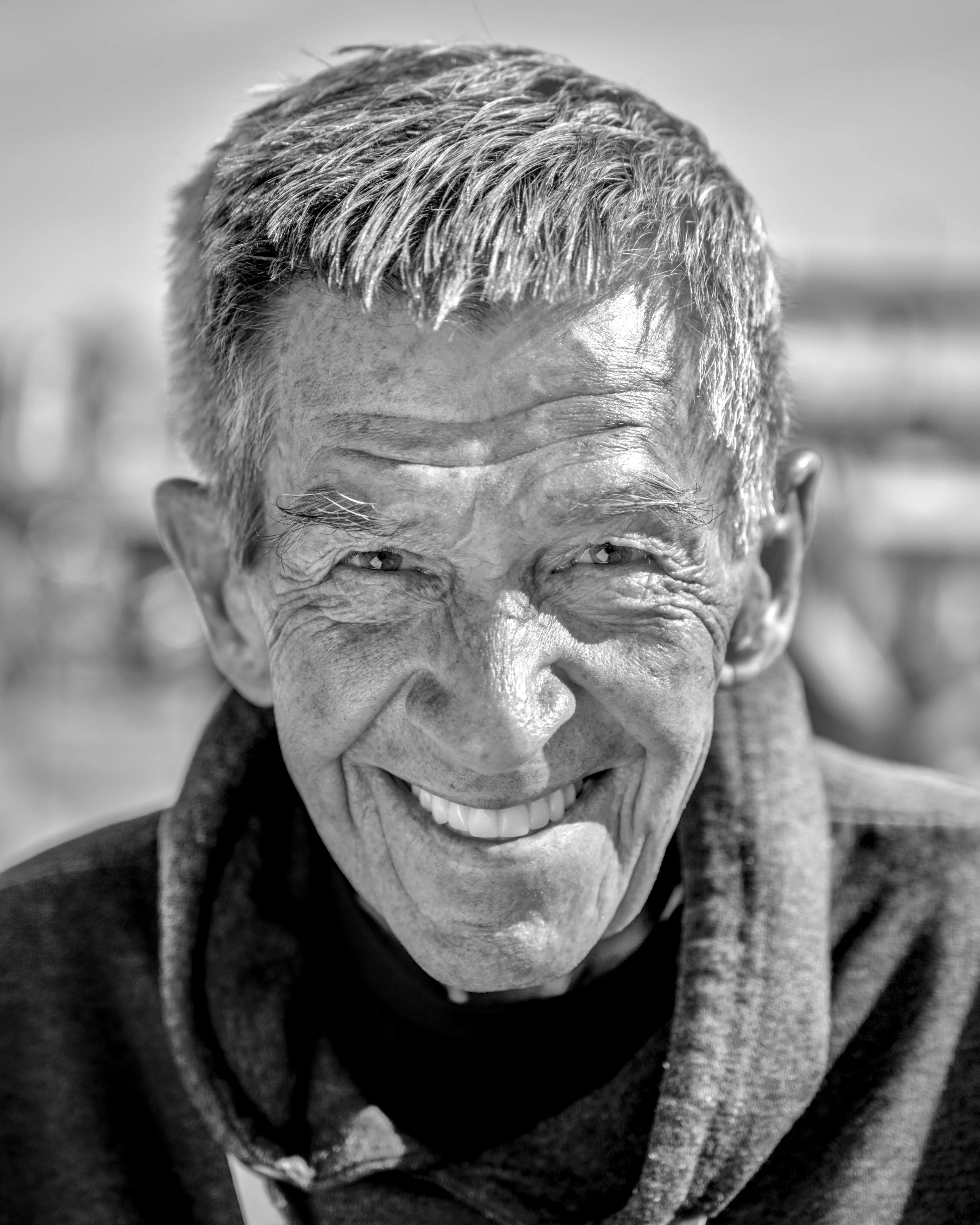 Close portrait of a smiling older person with short gray hair and a deeply lined face, wearing a dark hooded top.