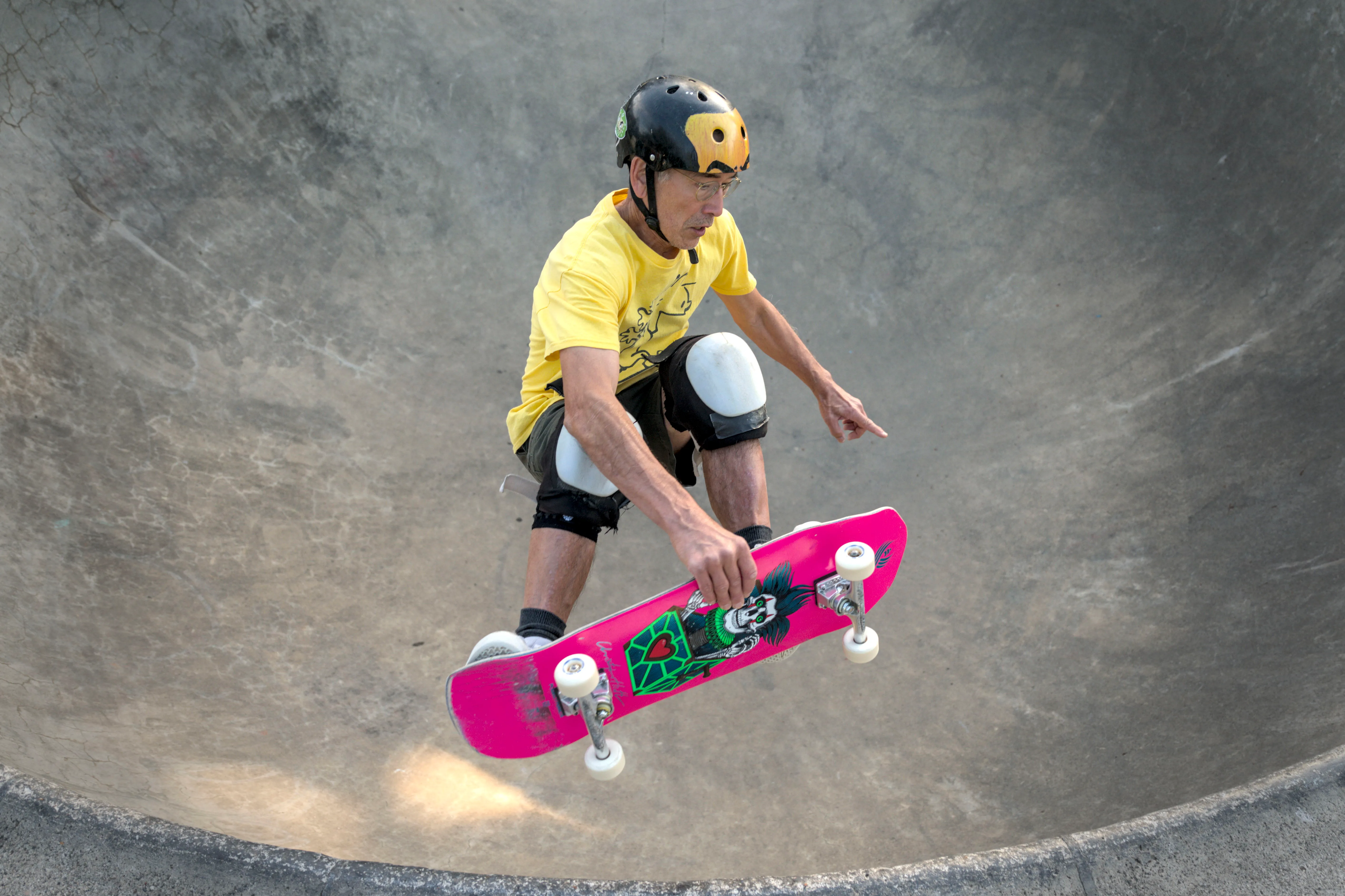 Skateboarder in a yellow shirt, helmet and knee pads airborne above a concrete bowl, holding a pink board.