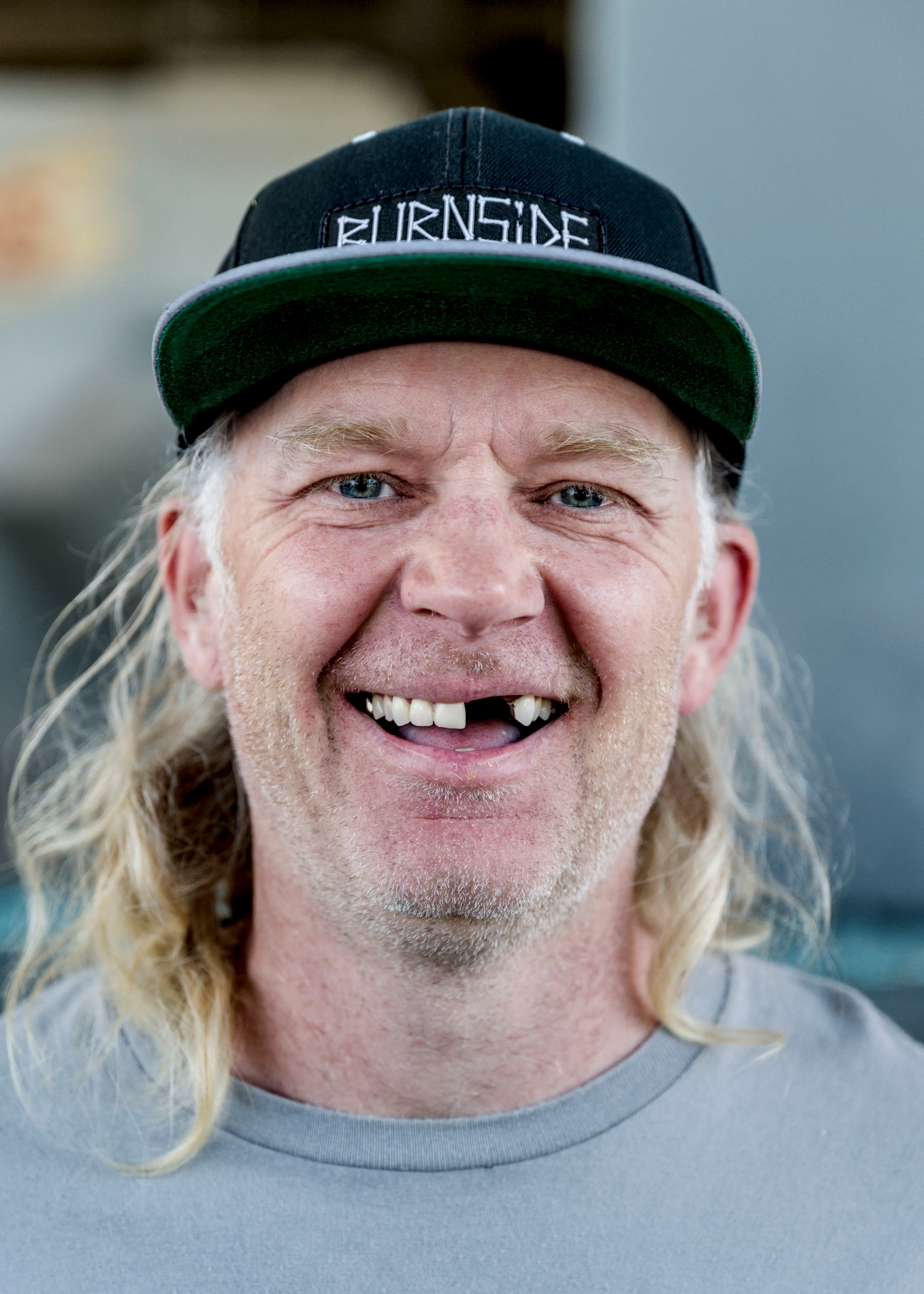 Smiling man with long blond hair, a black snapback cap and a gray t-shirt, missing a front tooth.