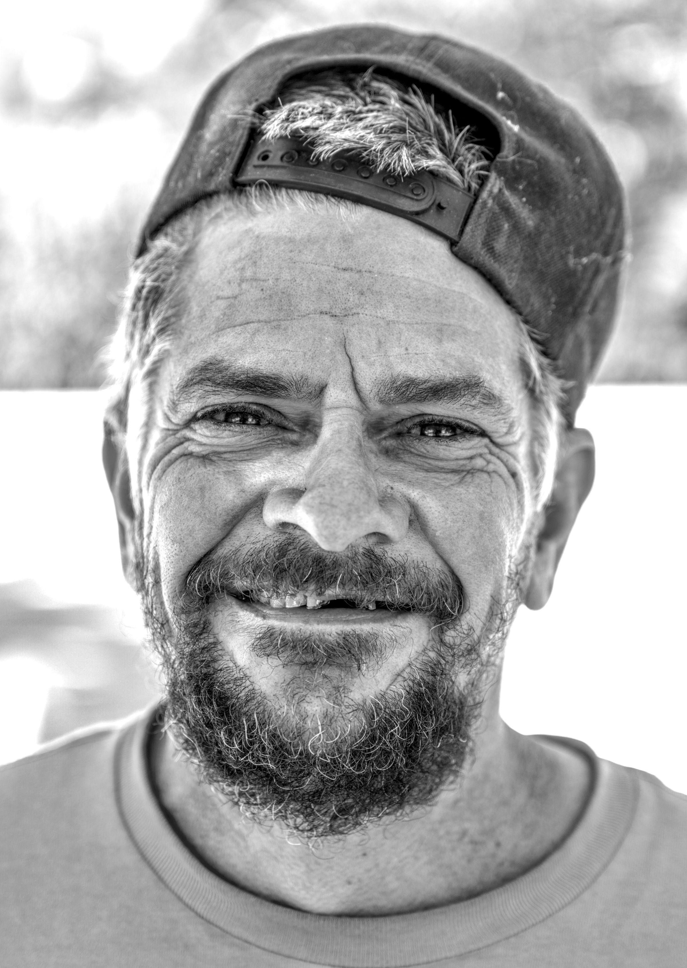 Close-up of a bearded man in a backward cap and t-shirt, smiling with creased eyes.