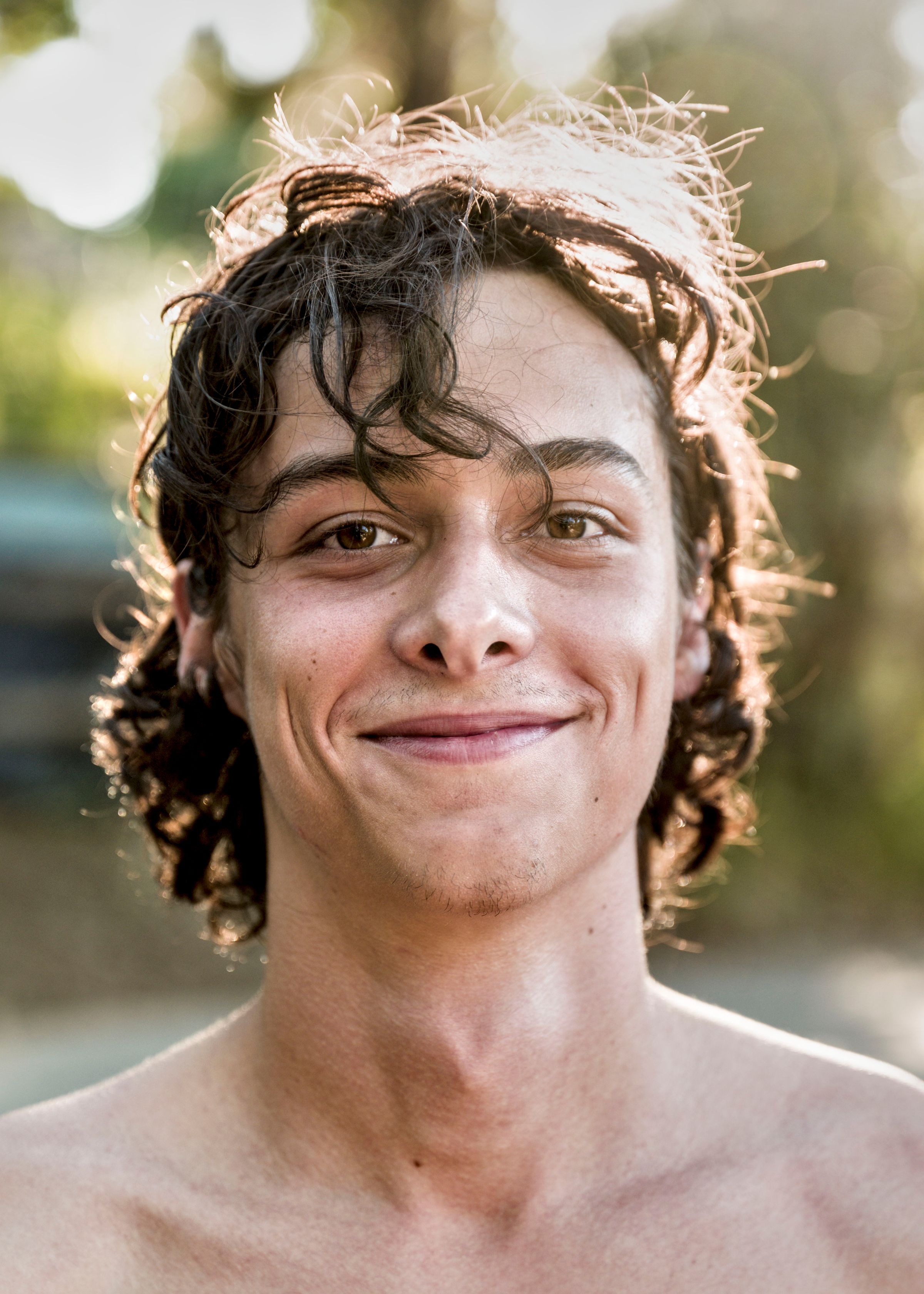 Shirtless young man with tousled curly dark hair lit from behind, smiling slightly at the camera outdoors.