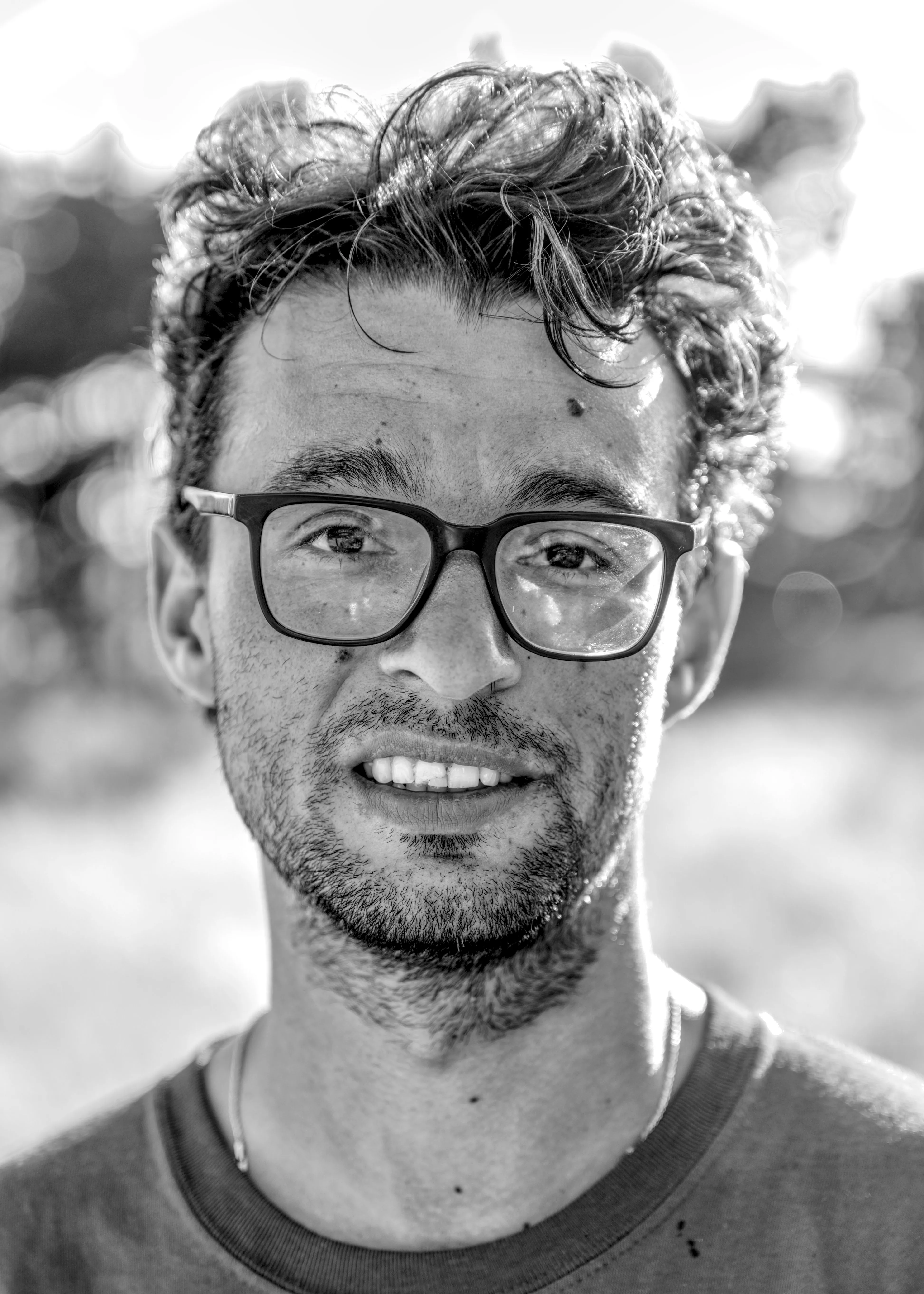 Close portrait of a smiling man with curly hair, dark-framed glasses and stubble, wearing a crew-neck T-shirt outdoors.