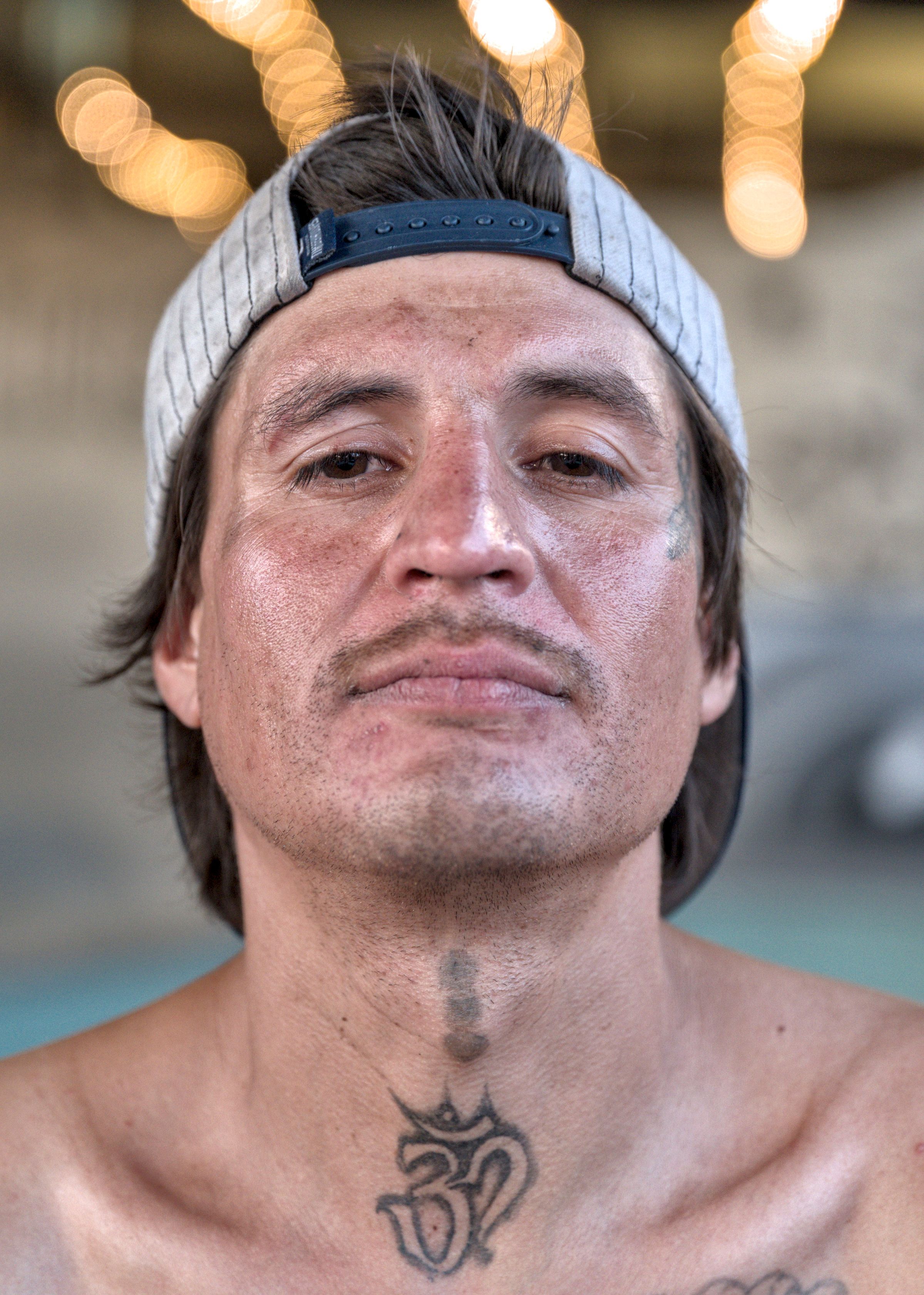 Close portrait of a shirtless man in a backwards gray striped cap, with neck tattoos, string lights blurred behind him.