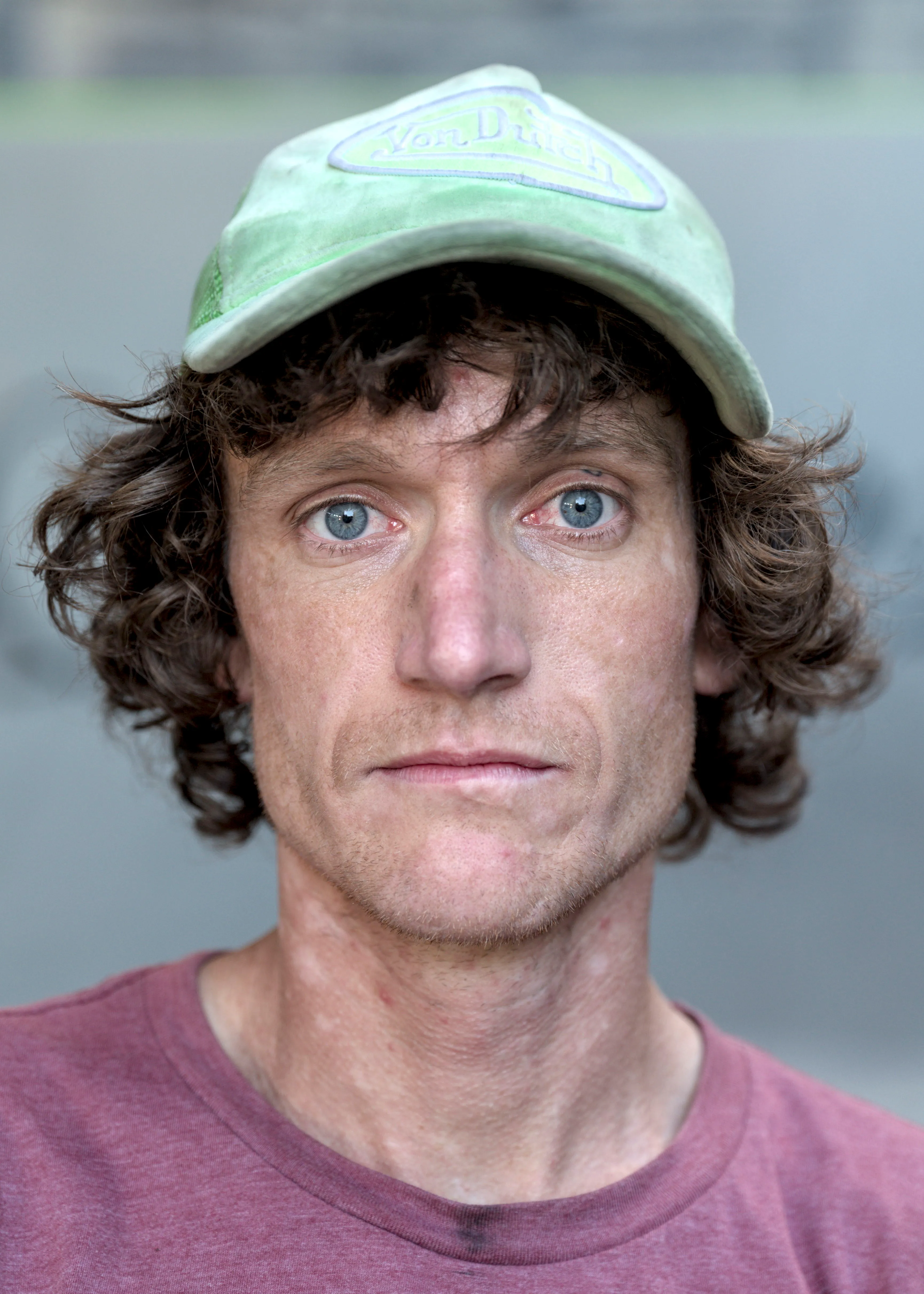 Close portrait of a man with curly brown hair, a green Von Dutch cap and a mauve t-shirt, looking at the camera.