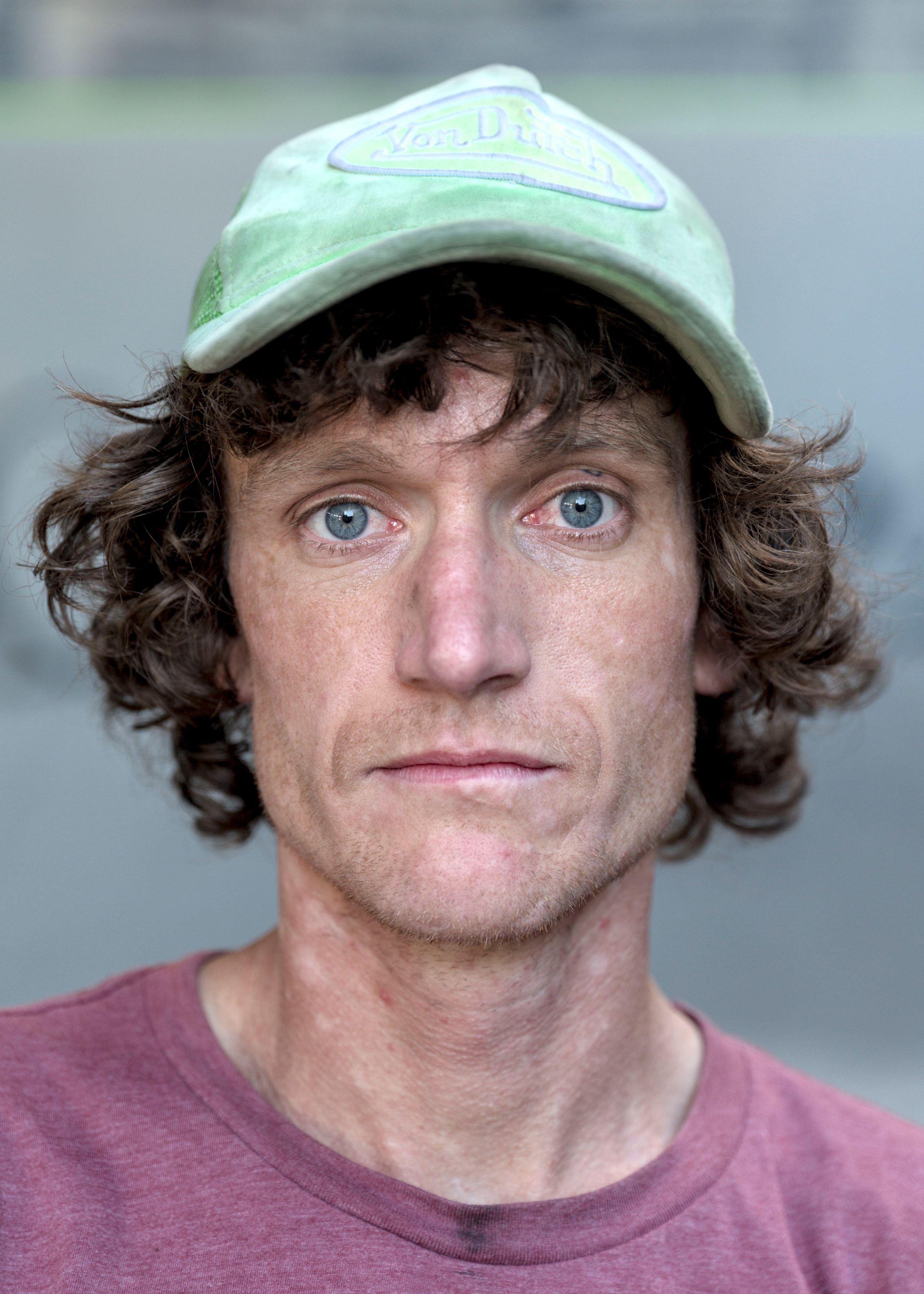 Close portrait of a man with curly brown hair, a green Von Dutch cap and a mauve t-shirt, looking at the camera.
