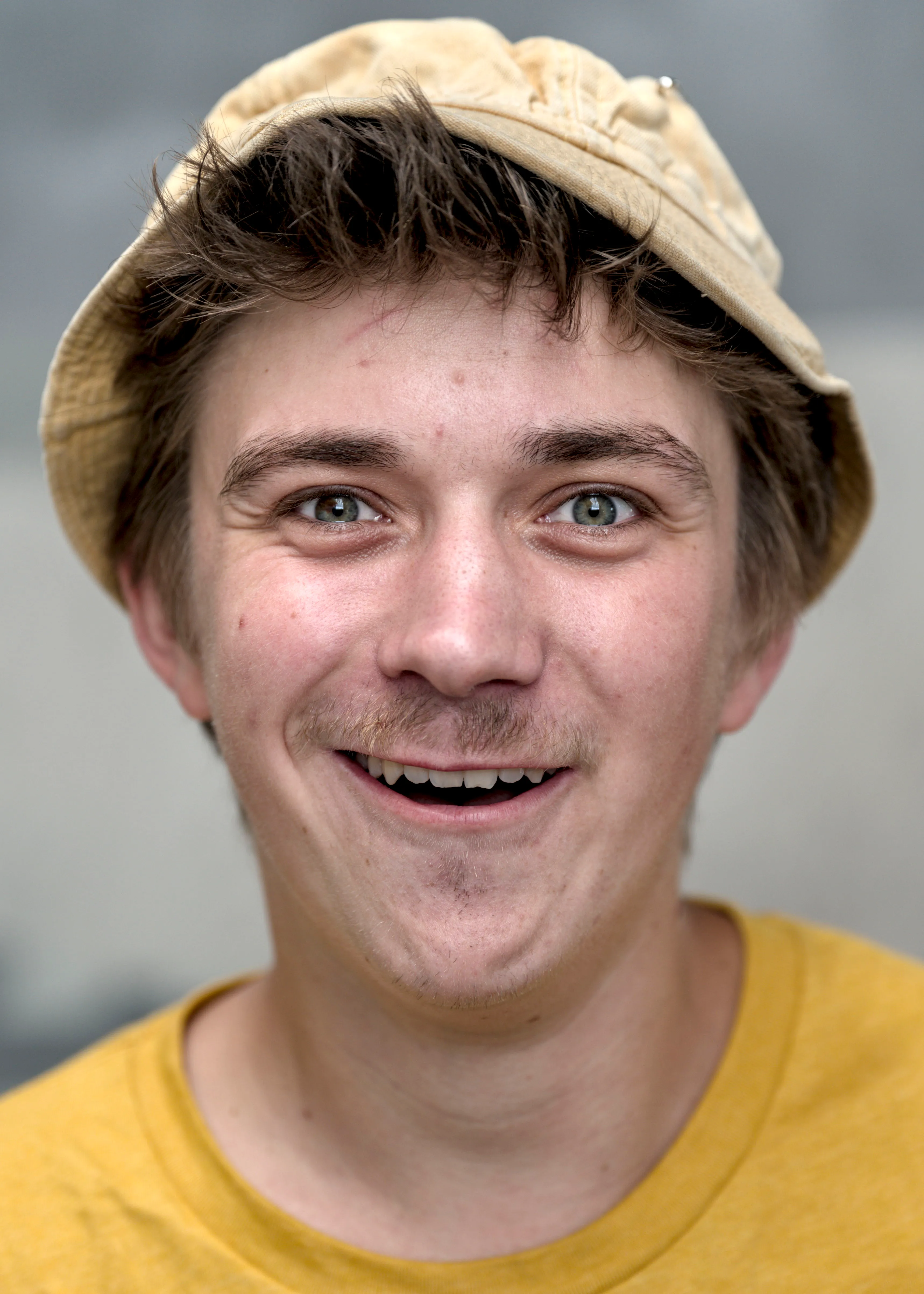 Close portrait of a smiling man in a tan bucket hat and mustard-yellow T-shirt, with a light mustache.