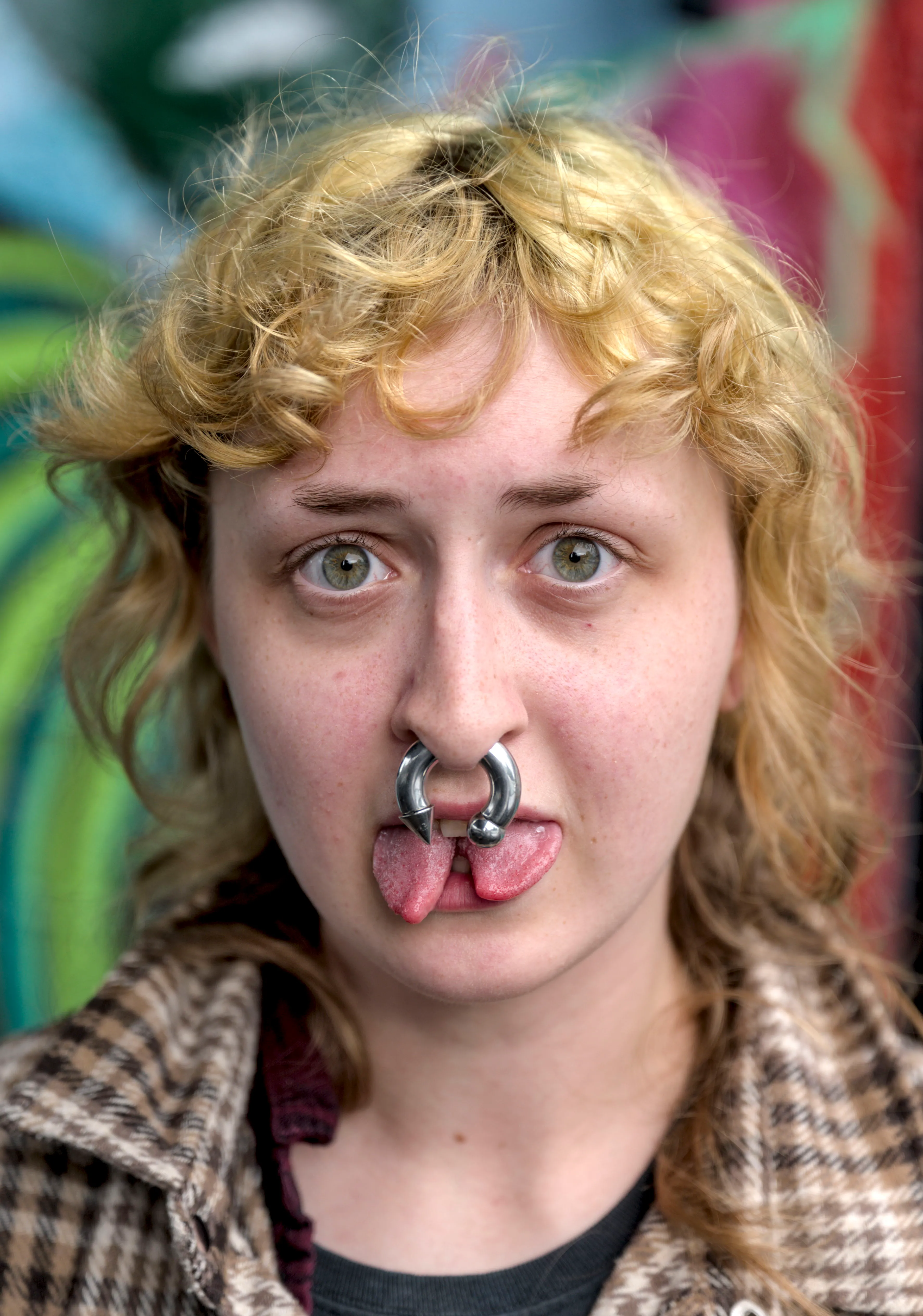 Close portrait of a person with curly blond hair, a large nose ring and a split tongue, in a plaid jacket before a colorful mural.