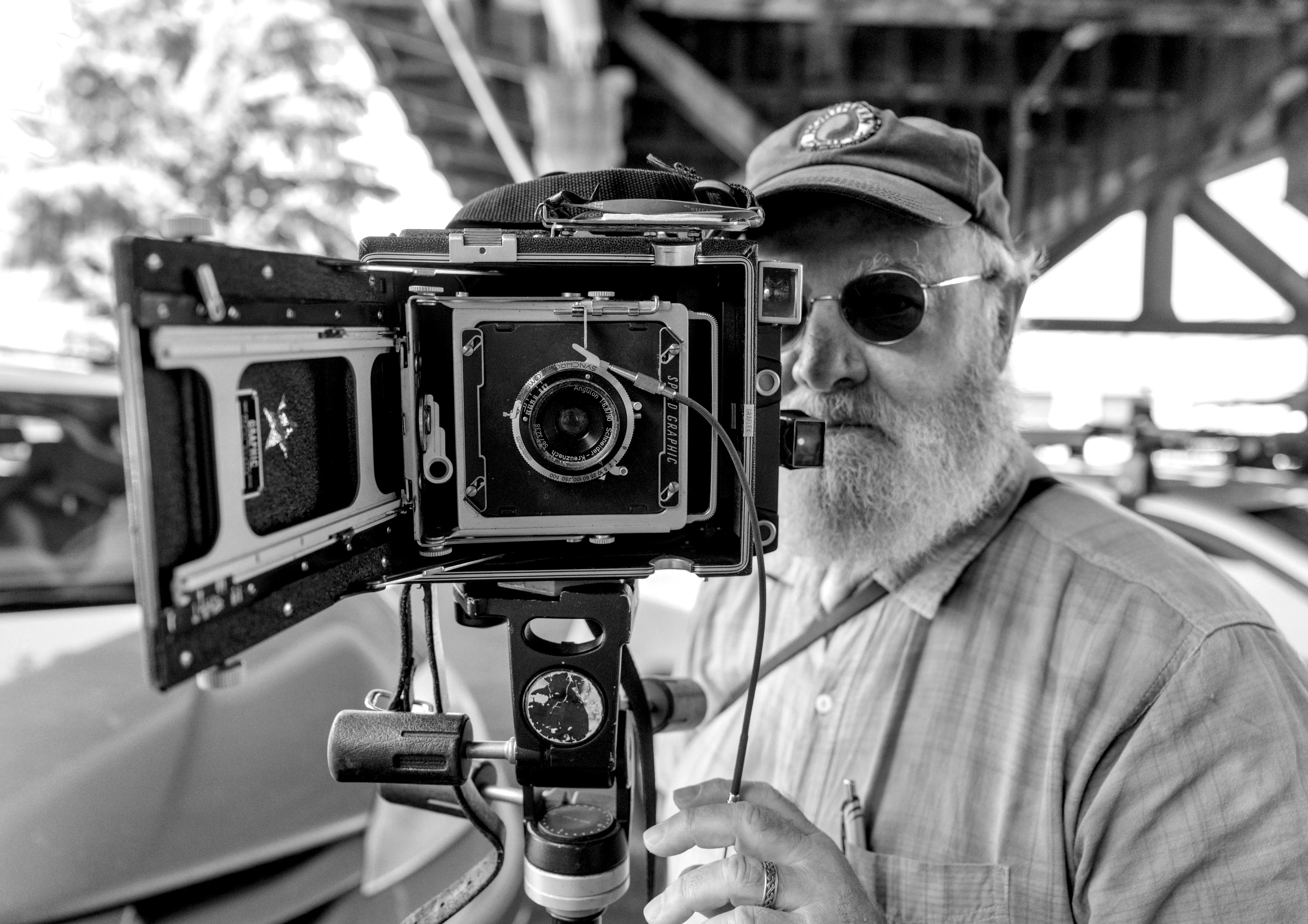 Bearded man in a cap and sunglasses behind a large-format field camera on a tripod, under a bridge.