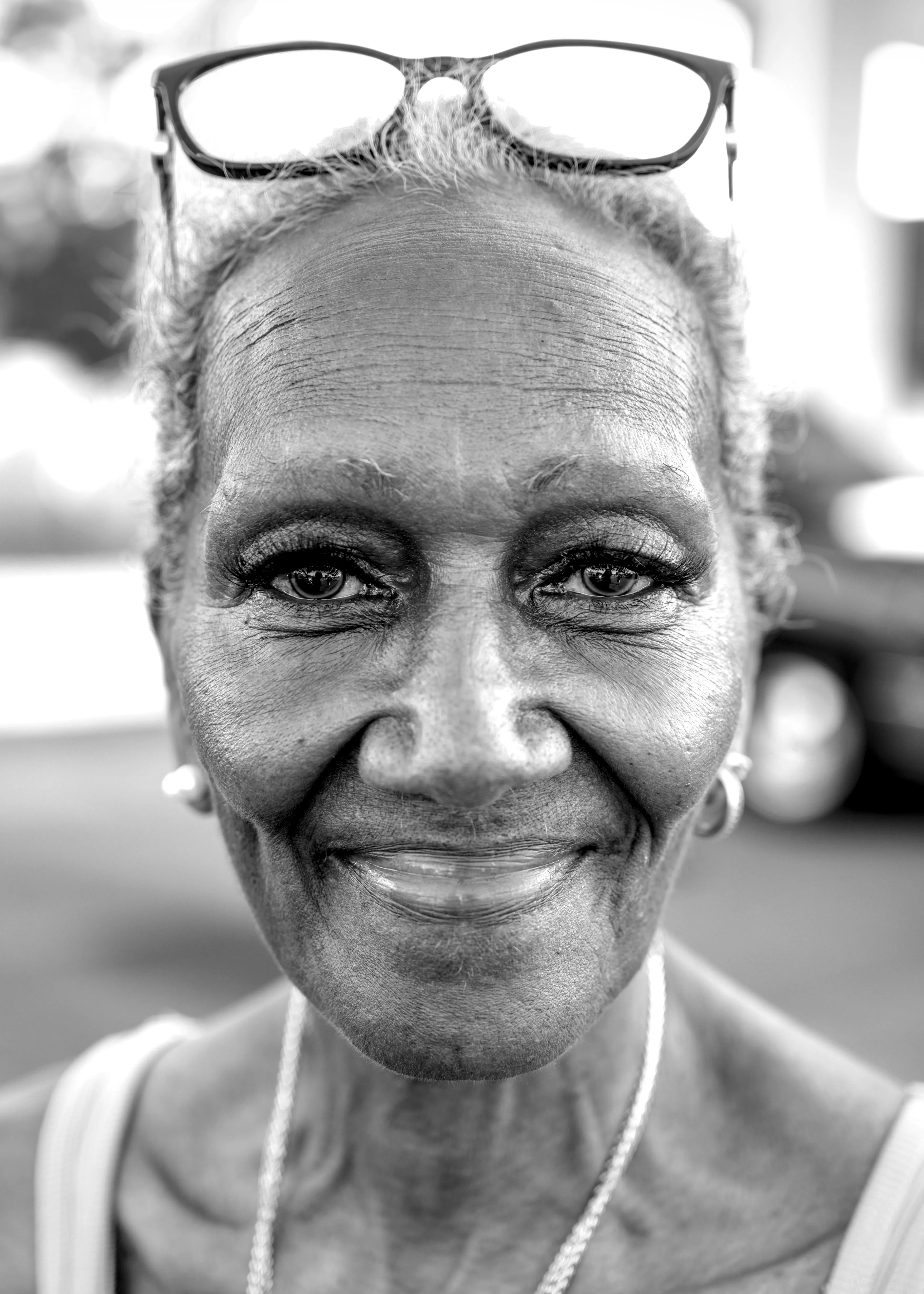 Close portrait of a smiling older woman with glasses on her head, earrings and a chain necklace.