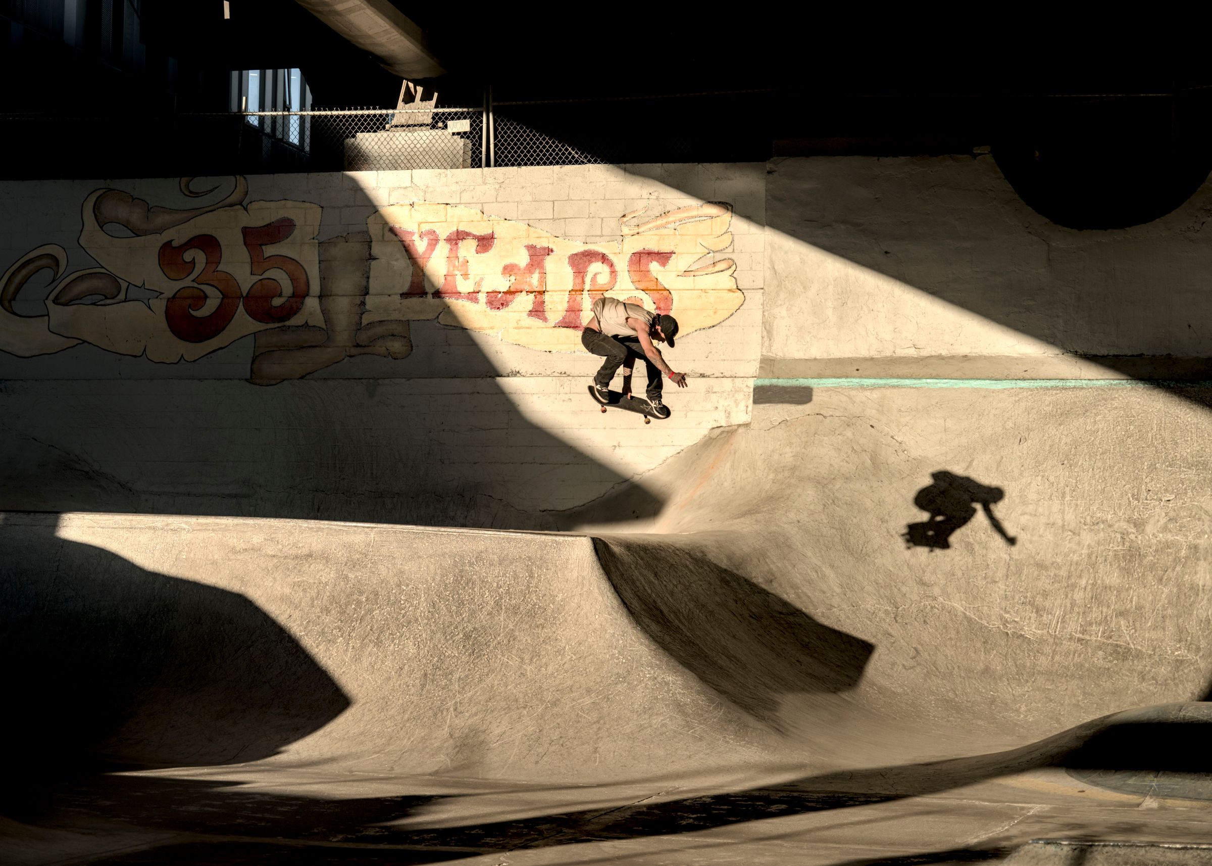 Skateboarder in midair above a concrete bowl, lit by a shaft of sun, beneath a painted "35 Years" sign on a brick wall.