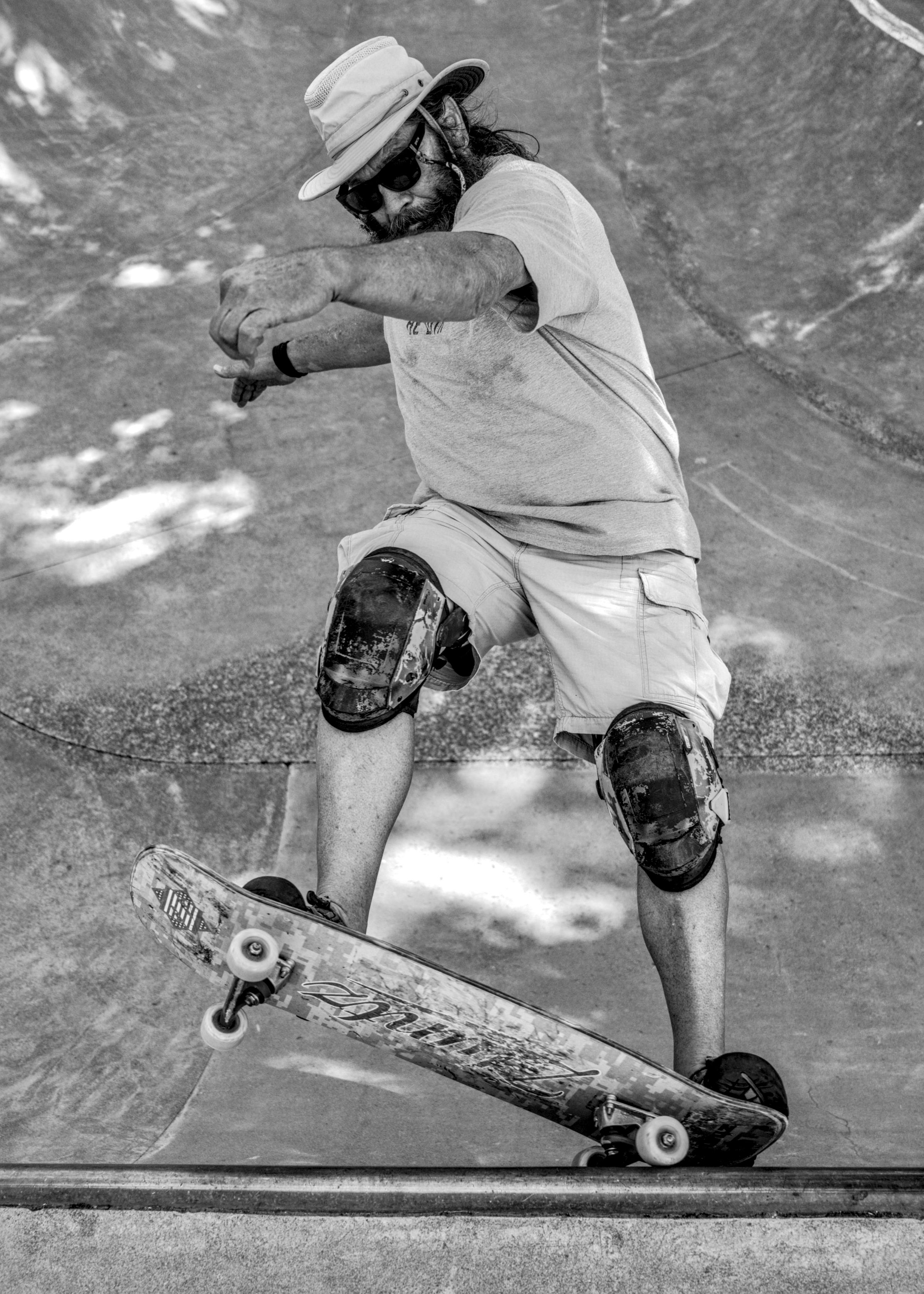 Bearded man in a brimmed hat, sunglasses and knee pads rides a tilted skateboard at the edge of a concrete bowl.