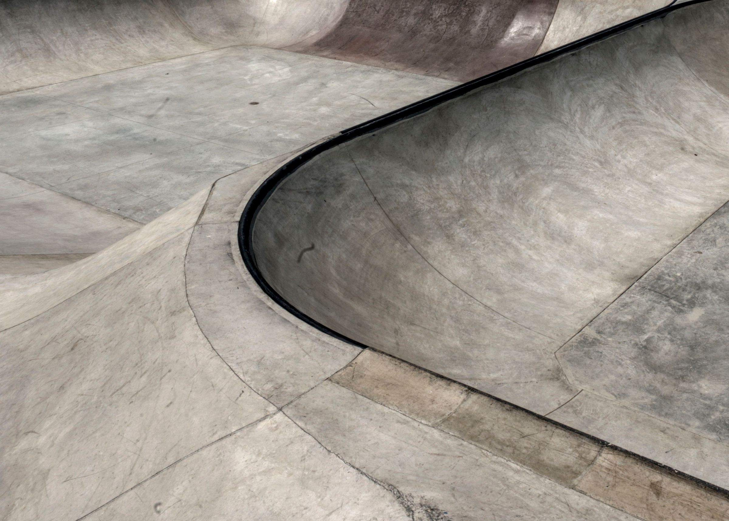 Curved concrete bowl edge with black metal coping and scuffed, worn surfaces at a skatepark.