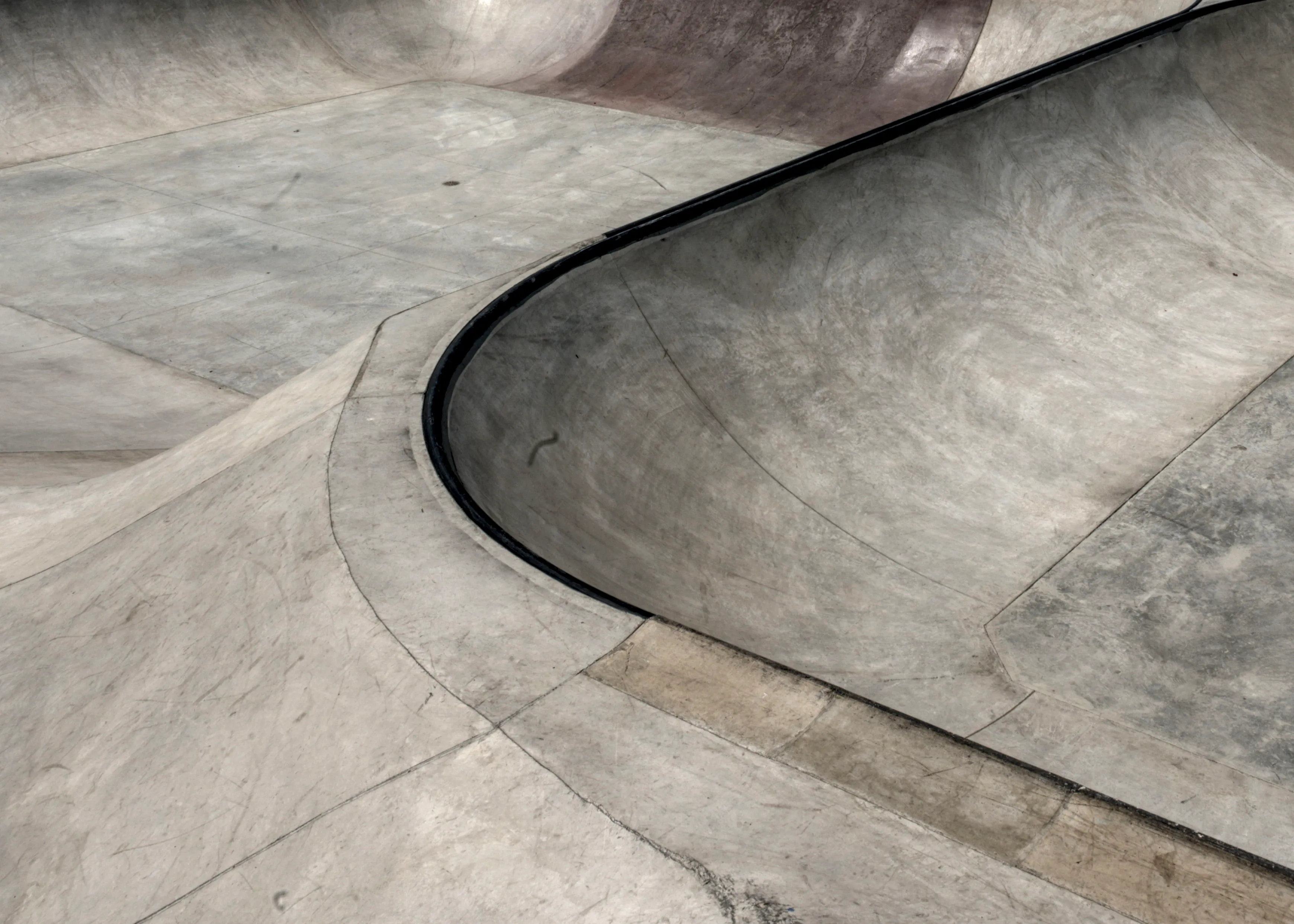 Curved concrete bowl edge with black metal coping and scuffed, worn surfaces at a skatepark.