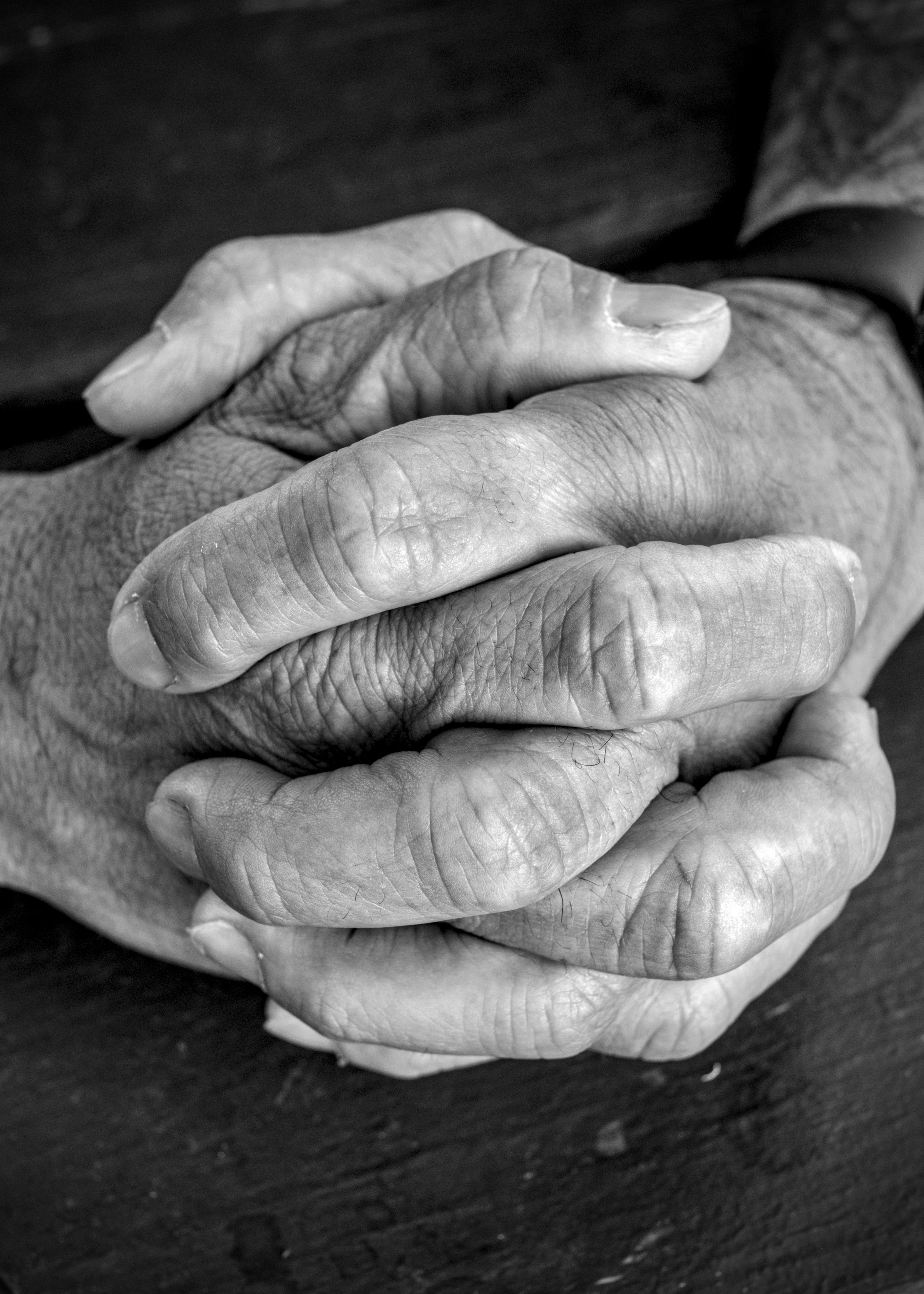 Close-up of two weathered hands clasped together with interlaced fingers on a dark wooden surface.