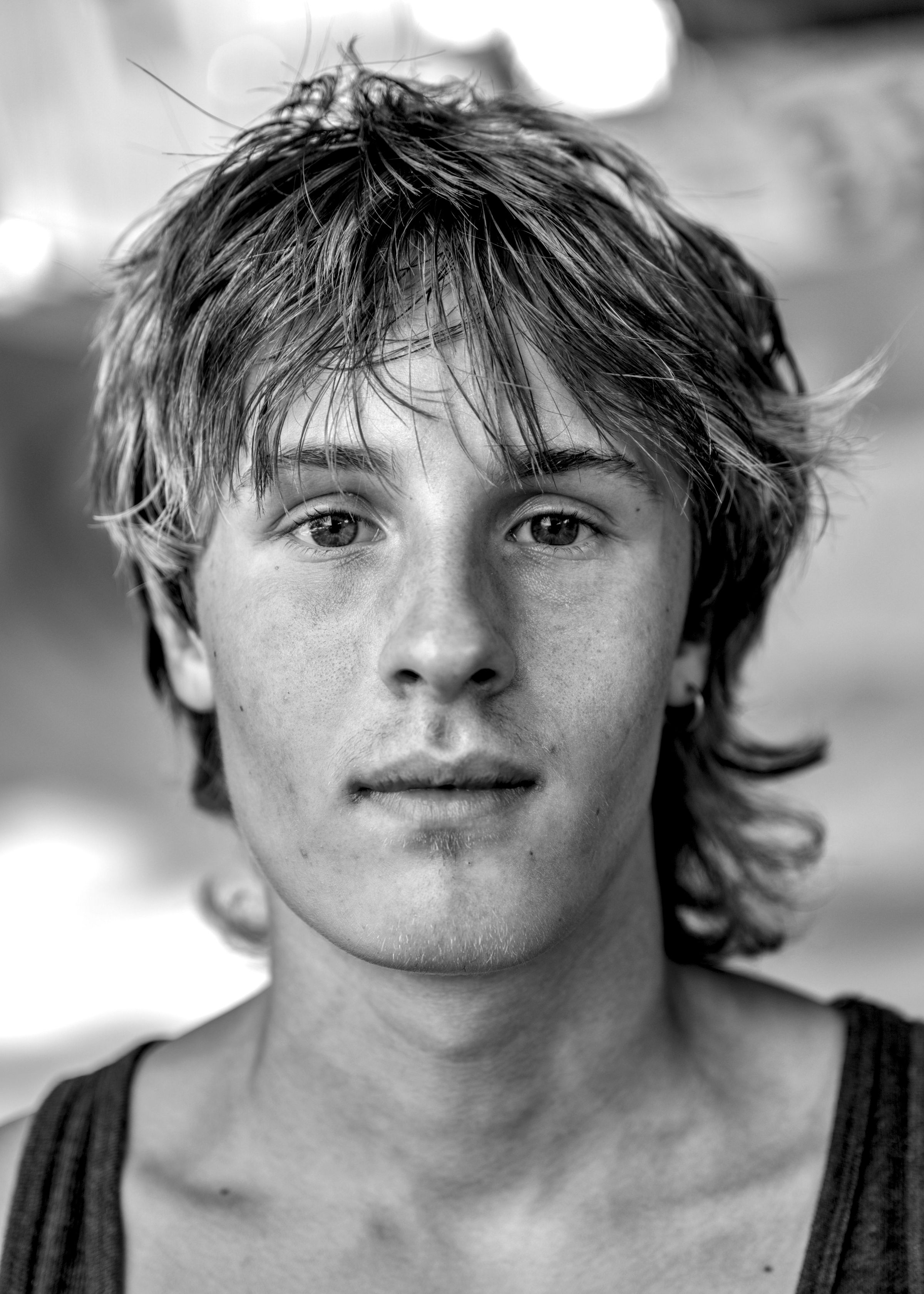 Close-up face of a young person with tousled shoulder-length hair, wearing a sleeveless top, against a blurred background.