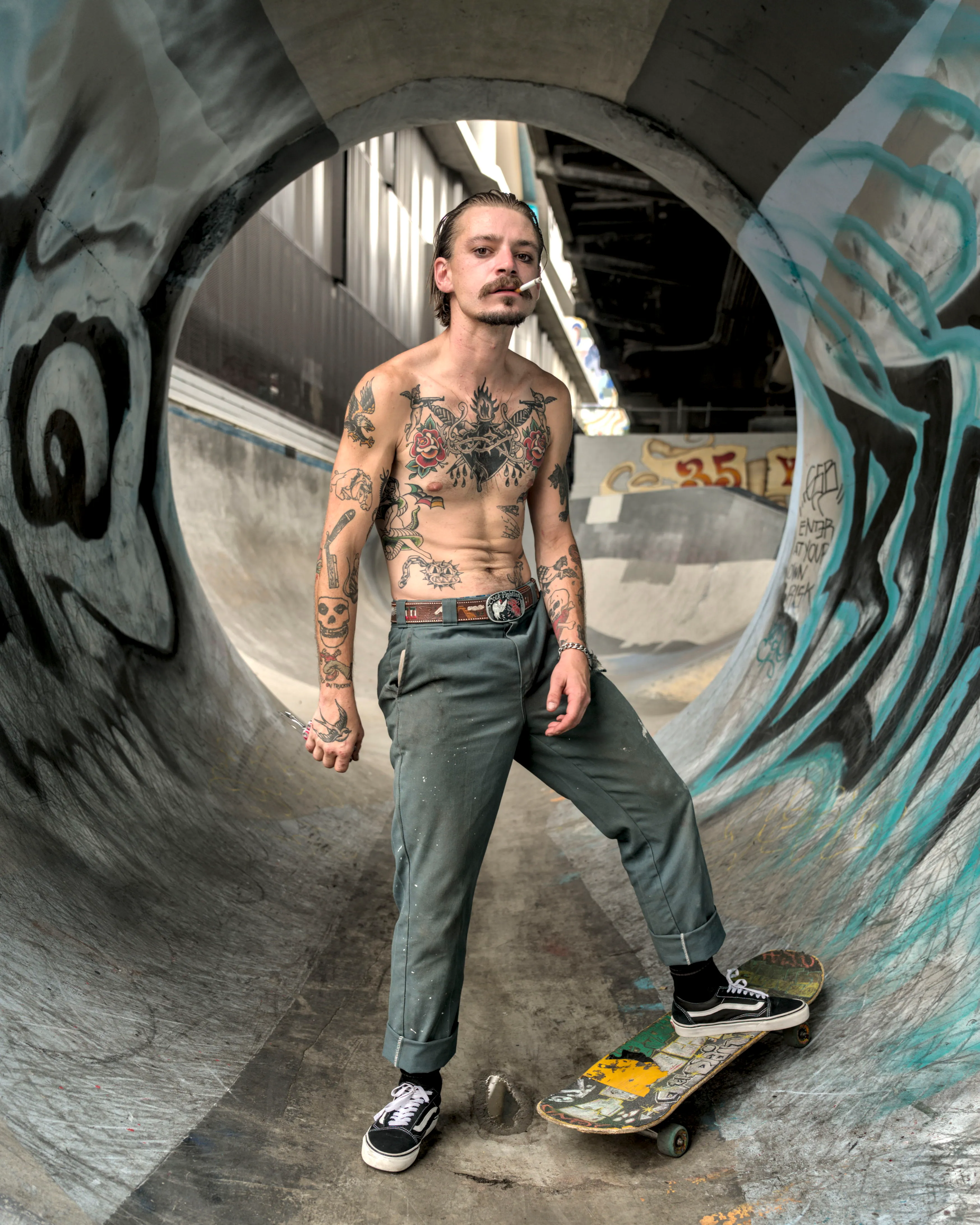 Shirtless tattooed man with a cigarette in his mouth stands in a graffitied concrete pipe, one foot on a skateboard.