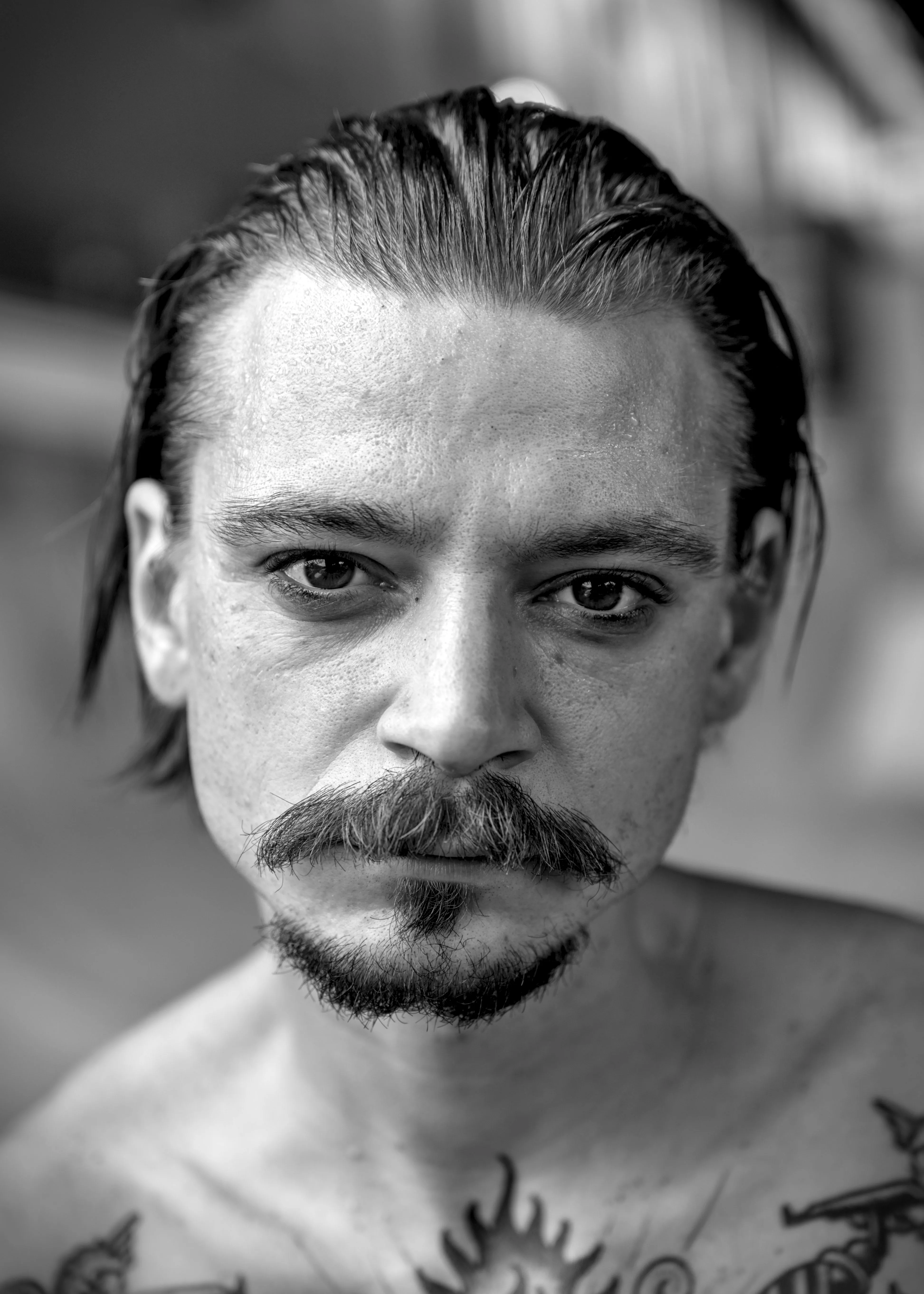 Close-up face of a shirtless man with slicked-back hair, a mustache and goatee, and tattoos on his chest.