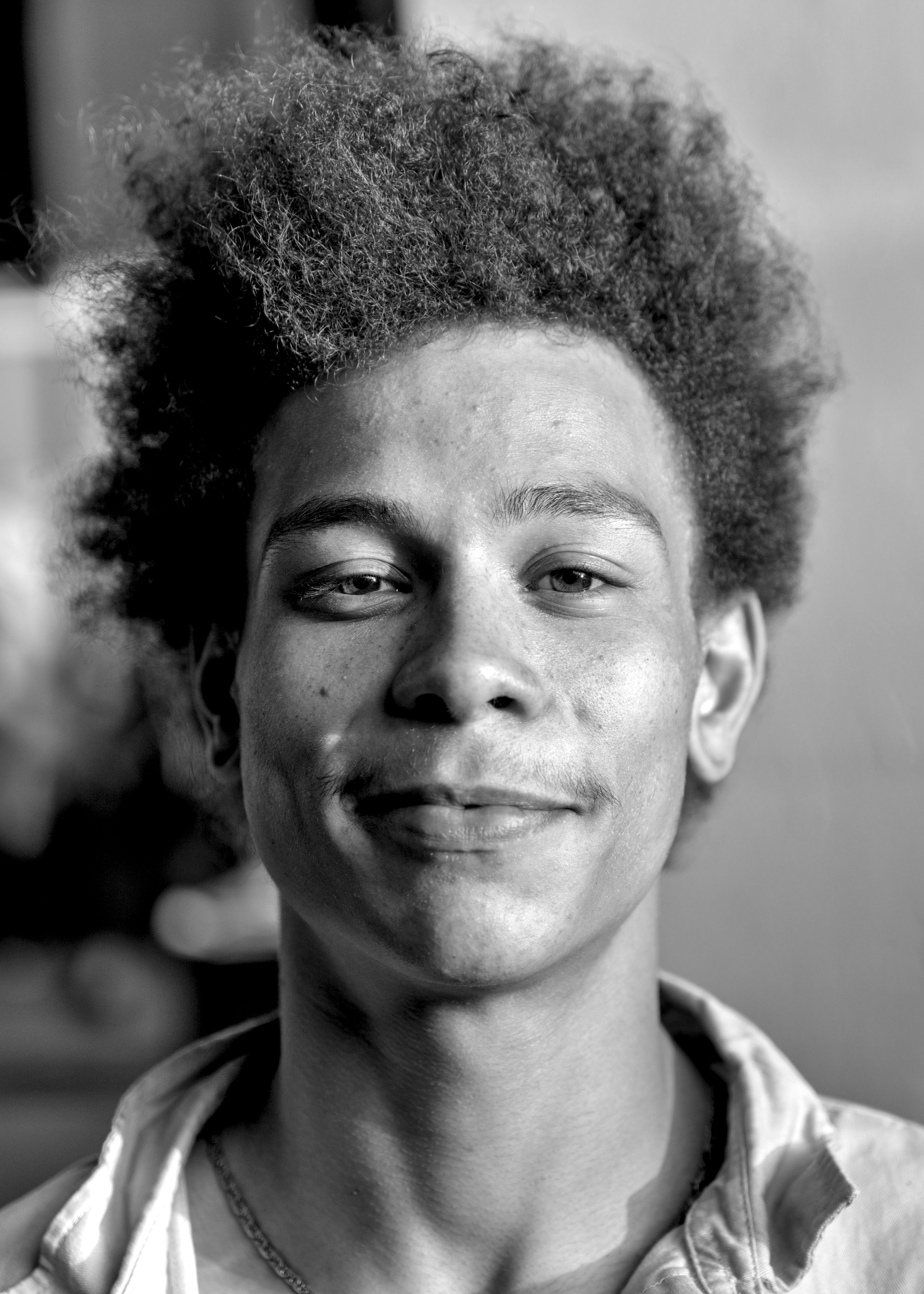 Close-up of a smiling young man with voluminous curly hair, wearing a collared shirt and a thin chain necklace.