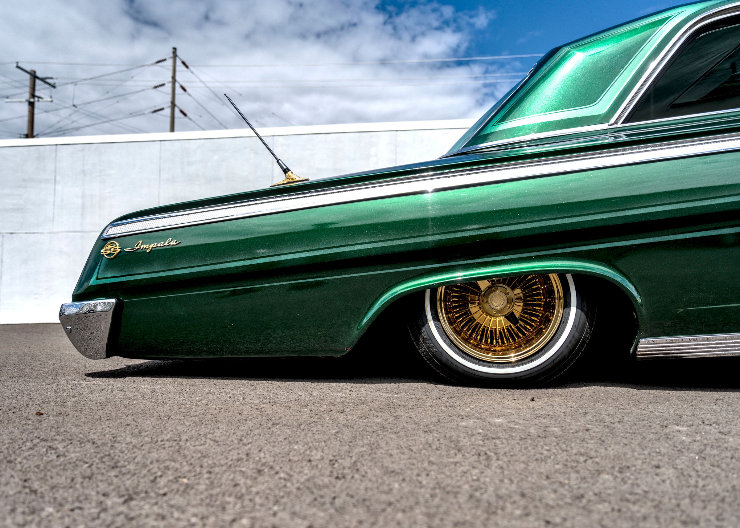 Front fender and gold wire wheel of a lowered green Chevrolet Impala parked on asphalt in front of a white wall.