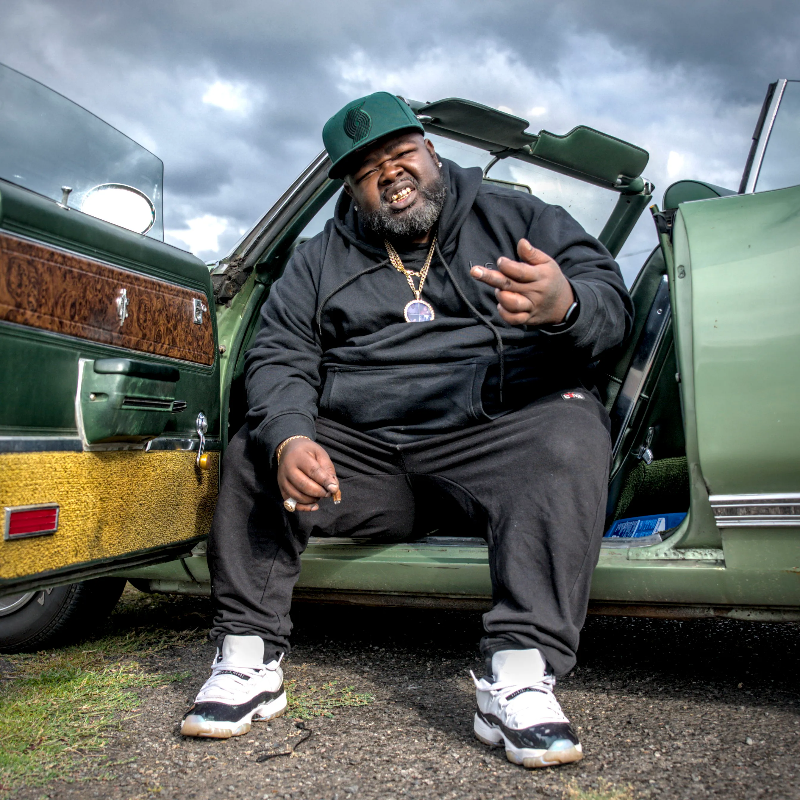 Man in a green cap and black hoodie sits in the open door of a green convertible, under a cloudy sky.
