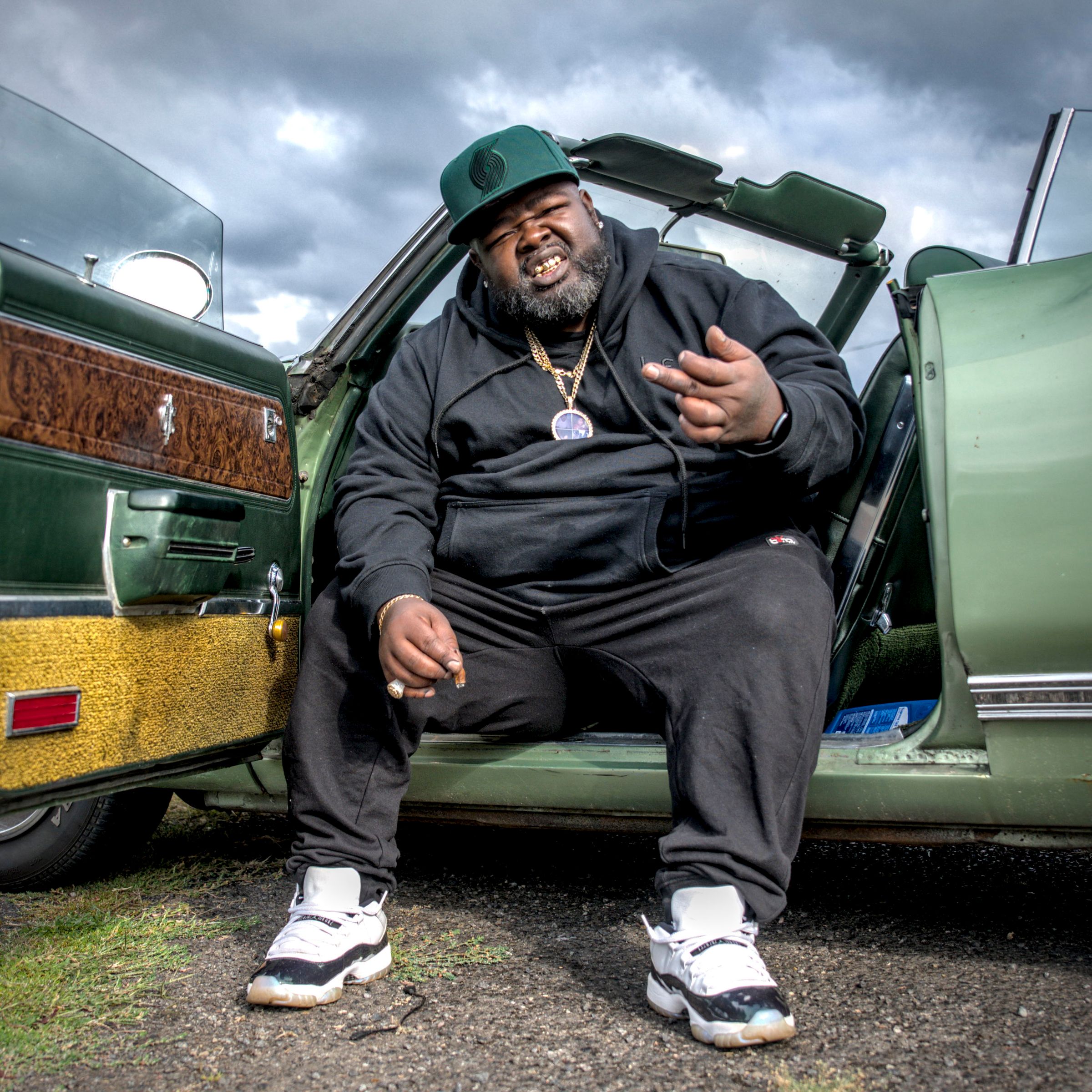 Man in a green cap and black hoodie sits in the open door of a green convertible, under a cloudy sky.