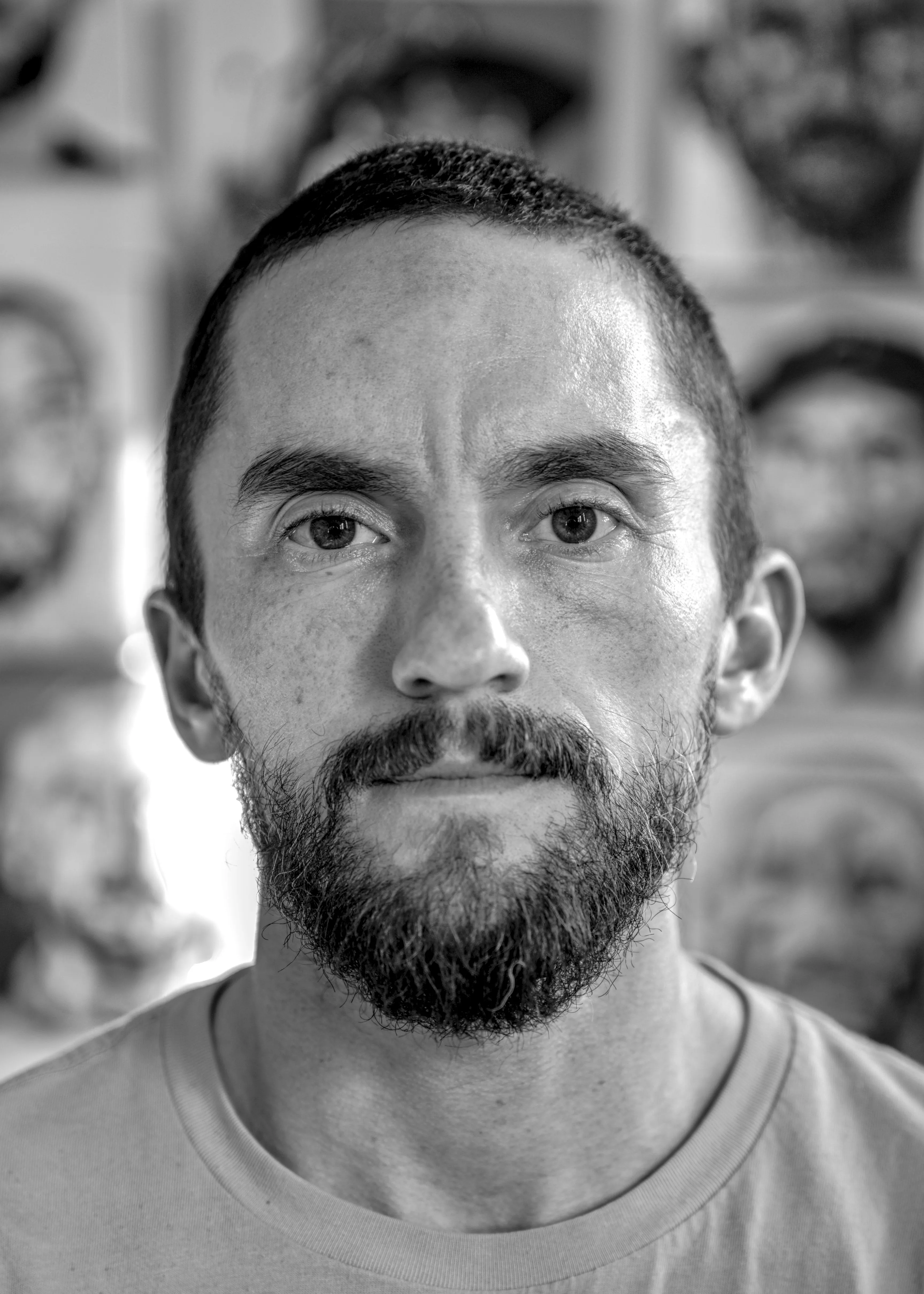 Close-up of Tyler Bingham, a bearded man with close-cropped hair, in front of a wall of his painted self-portraits.