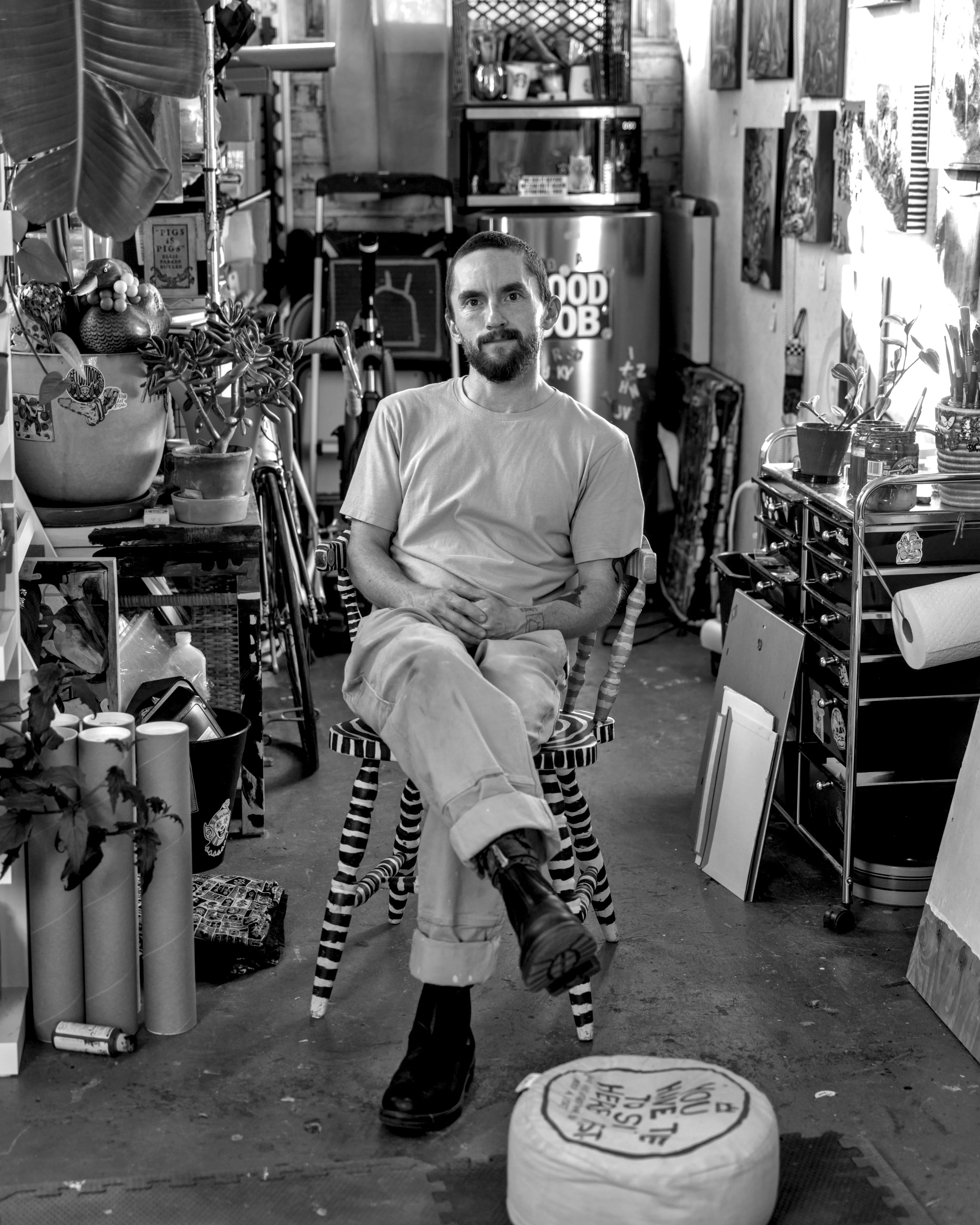 Tyler Bingham sits cross-legged on a striped wooden chair in his crowded art studio, surrounded by plants, paintings and supplies.